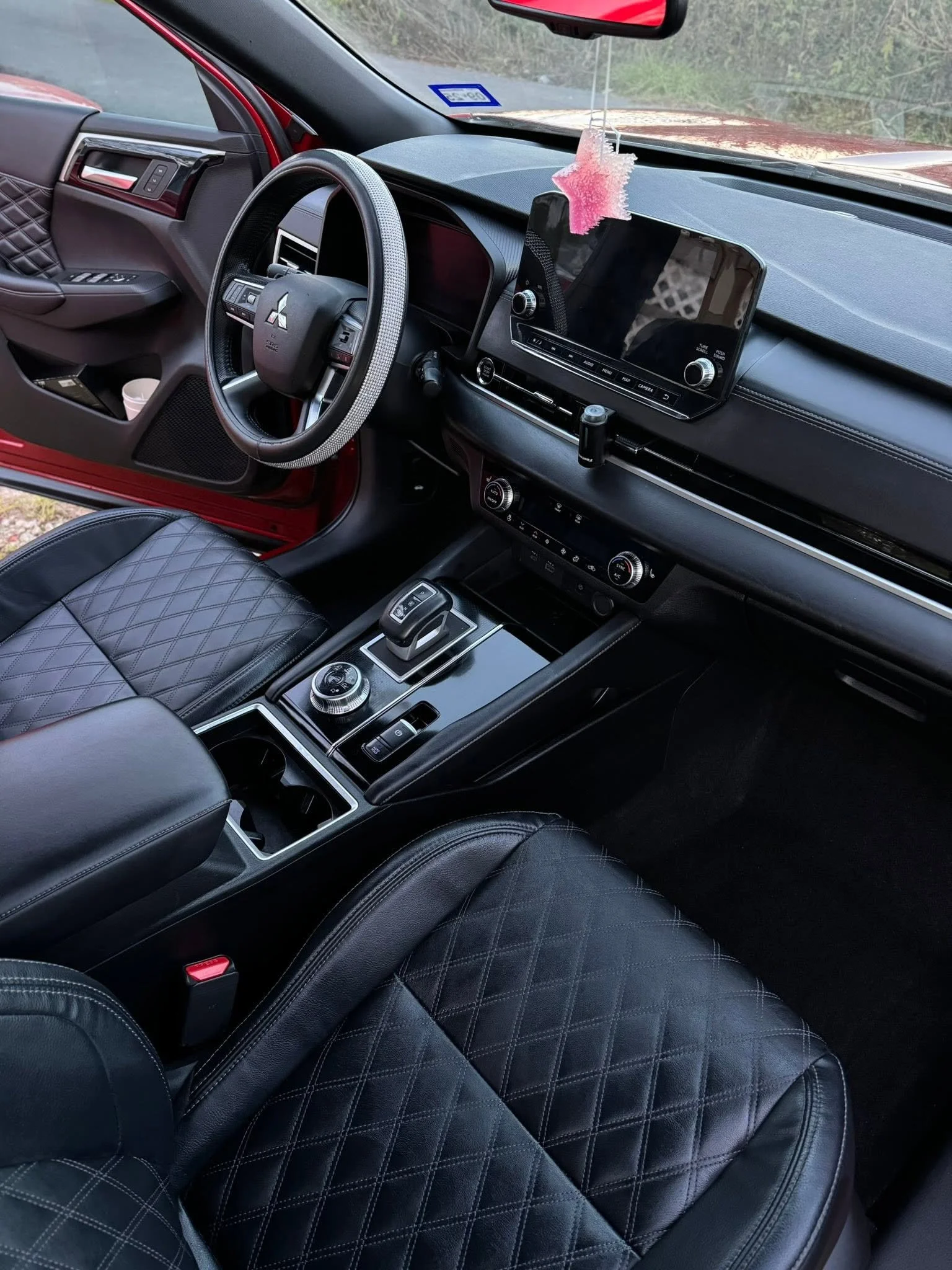 The interior of a car showing the front passenger and driver seats with black leather quilted upholstery, a dashboard with a digital touchscreen, steering wheel with controls, center console with gear shifter, and a pink decorative air freshener hang
