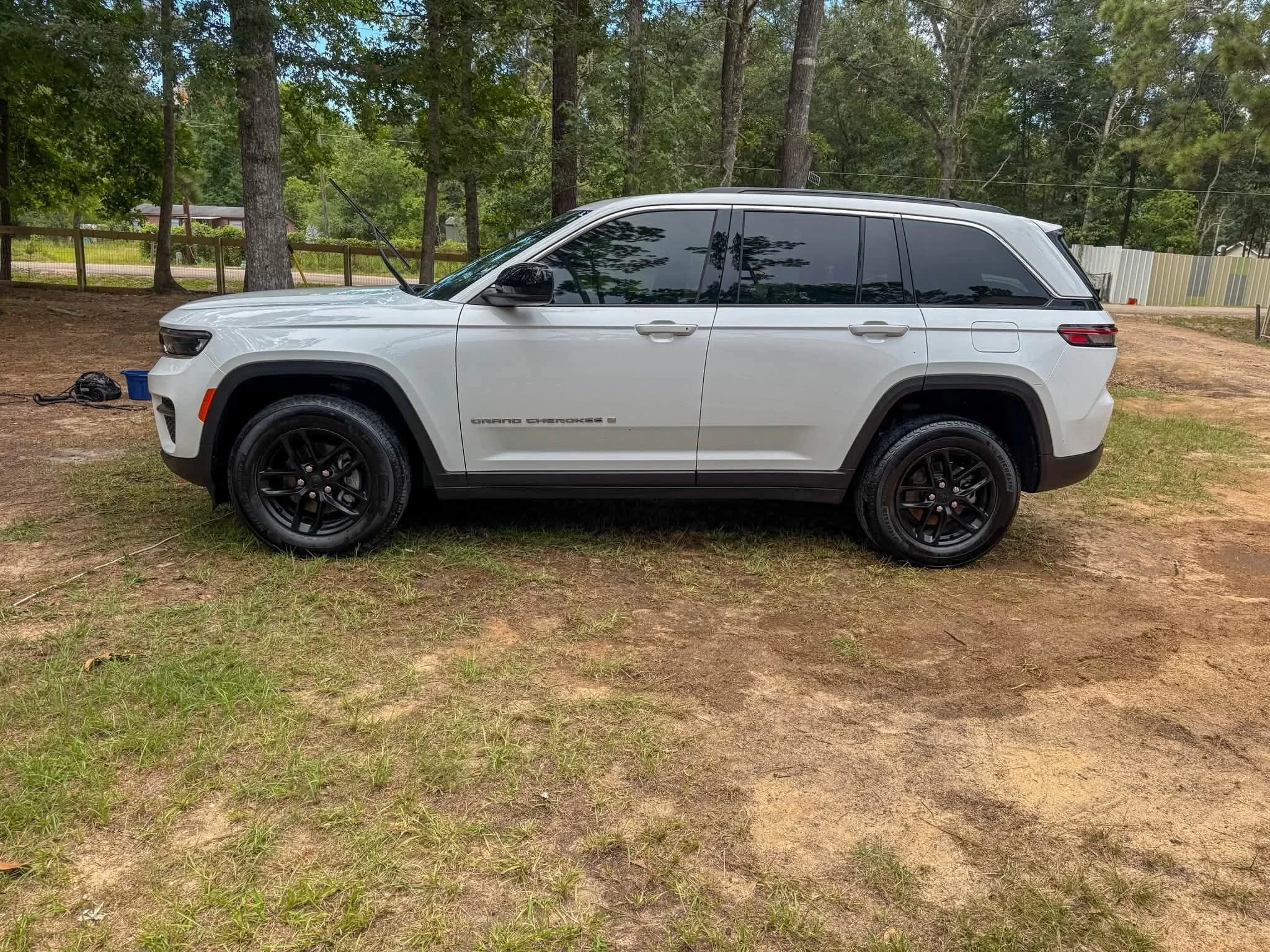 A white Jeep Grand Cherokee parked on a patch of dirt and grass in a wooded area with trees and a wooden fence in the background.