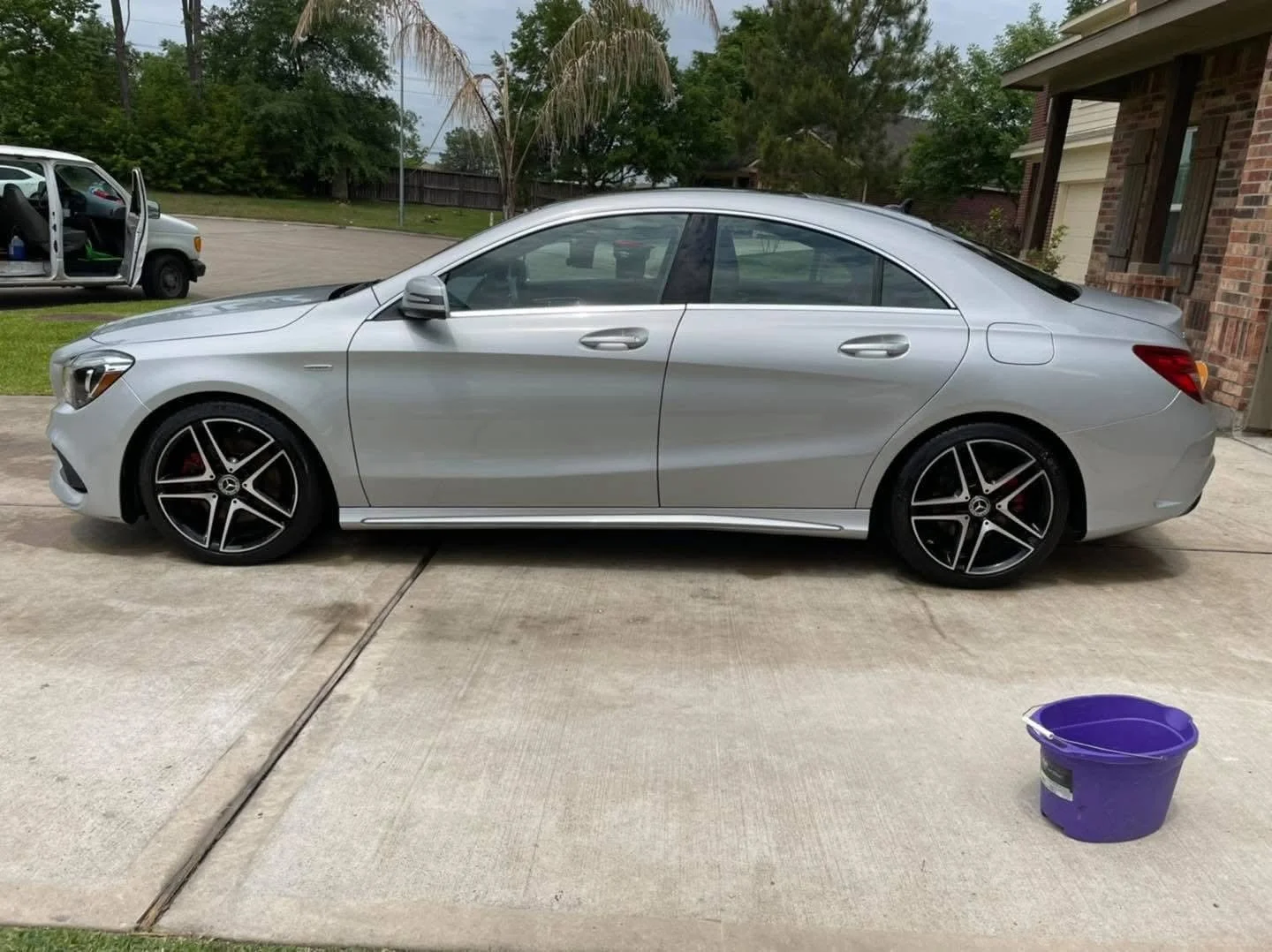 A silver luxury sedan parked on a driveway in a residential area, with a purple bucket in the foreground.