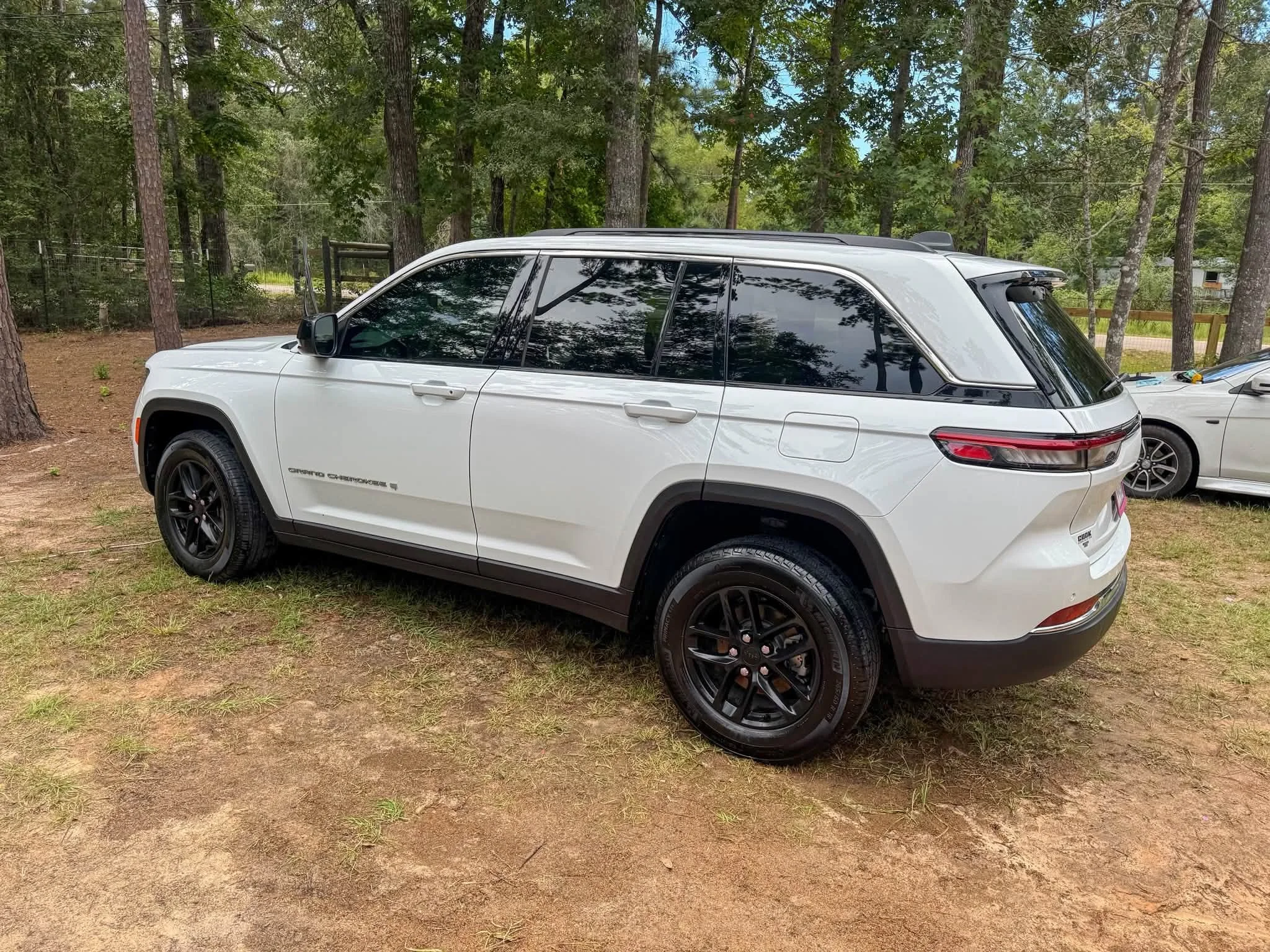 White Jeep Grand Cherokee parked on a grassy area surrounded by trees with a fence and a small building in the background.