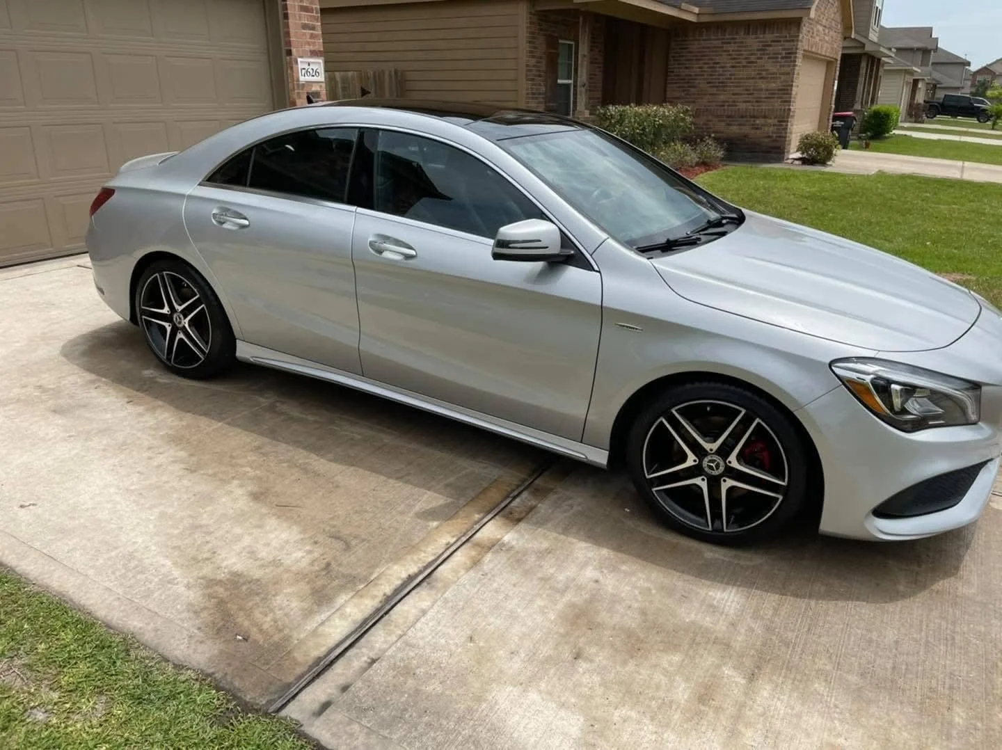 A silver luxury sedan parked in a residential driveway, with a brick house and garage in the background and green grass on the side.