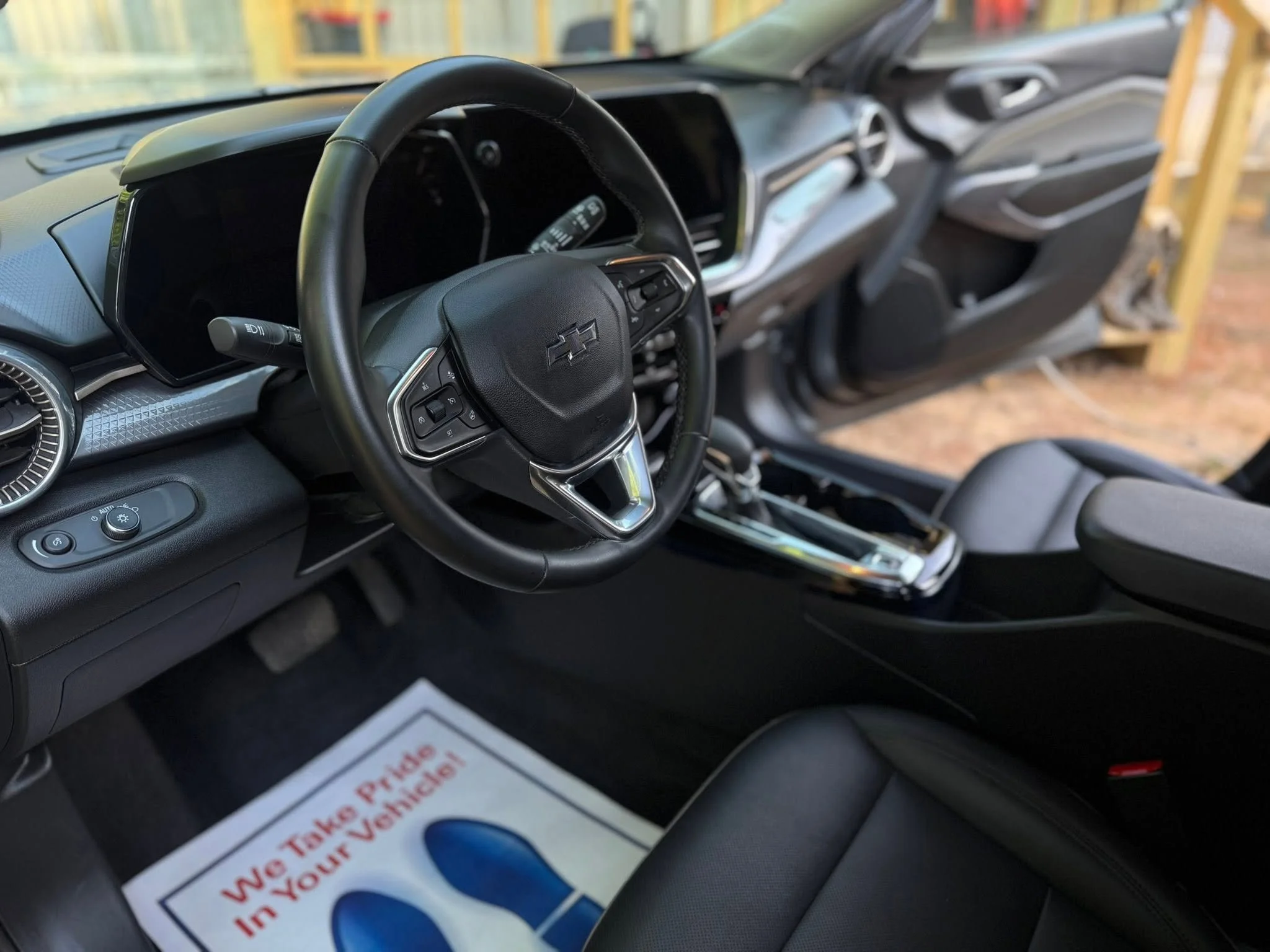 The interior of a vehicle showing a black steering wheel with car brand logo, dashboard, and a part of the front passenger area, with car seats and a window view outside.