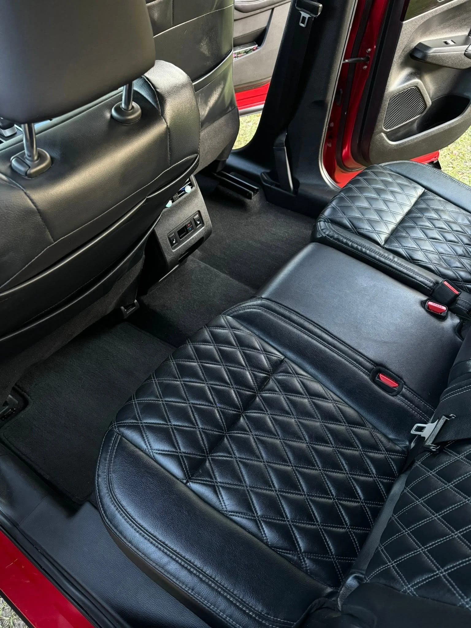 The interior of a vehicle showing black leather seats with diamond quilted stitching and black carpeted floor, with the open red door visible.