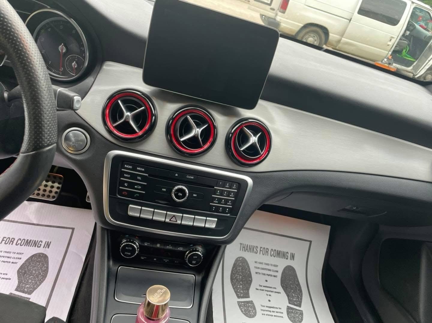 Interior of a car dashboard with three circular air vents with red accents, a central multimedia control panel, and a display screen above the vents. Part of the steering wheel and outside view of parked vehicles are visible.