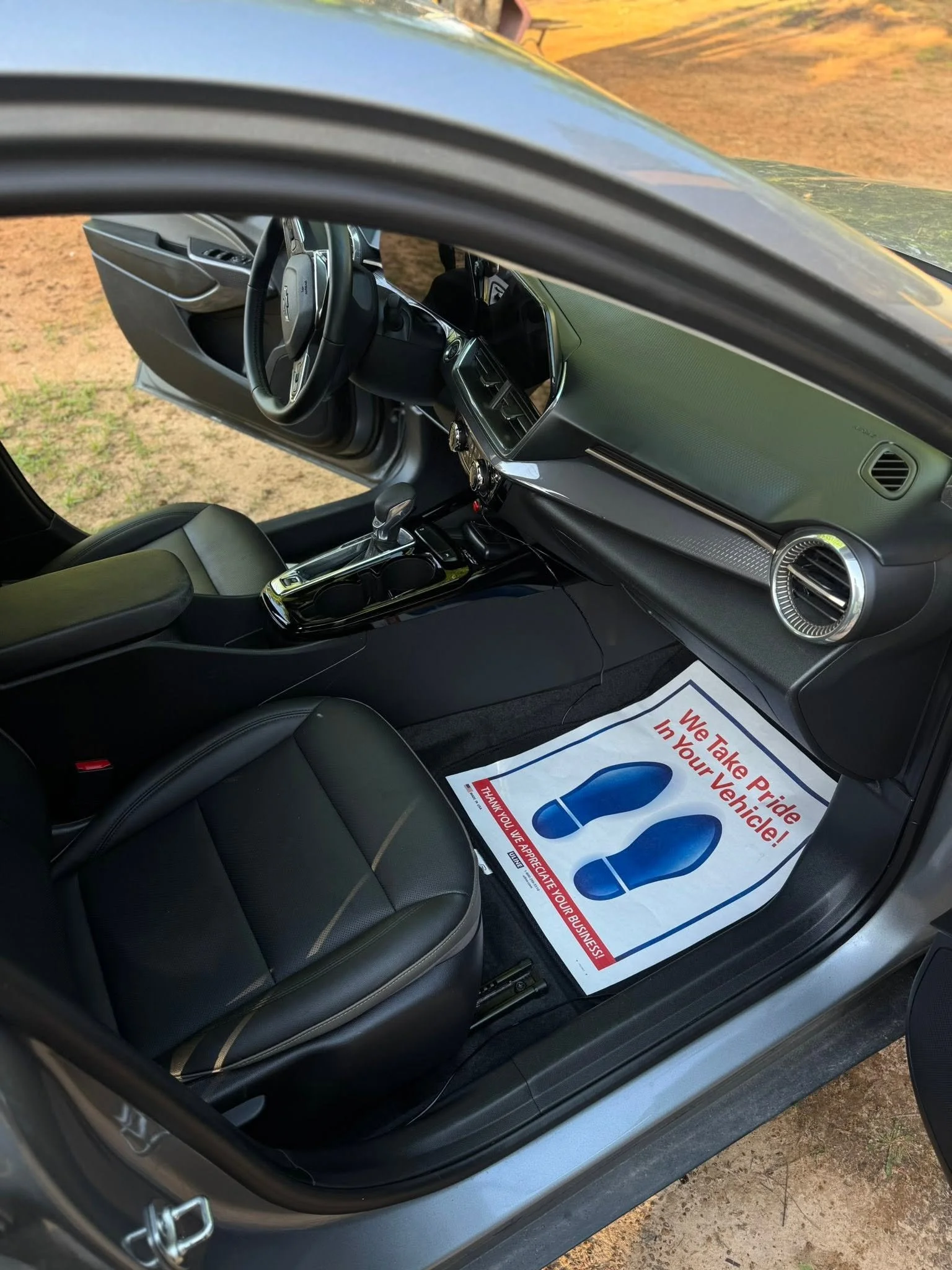 Interior of a car showing the driver's seat, steering wheel, dashboard, and a floor mat with a sign that reads, "We Take Pride In Your Vehicle!"