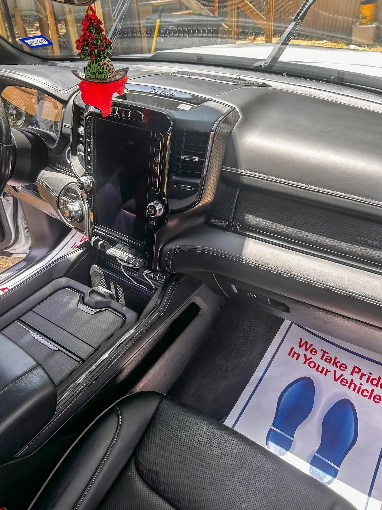 Inside a vehicle with a digital dashboard, a colorful air freshener hanging from the rearview mirror, and a sign on the floor that reads, "We Take Pride In Your Vehicle."