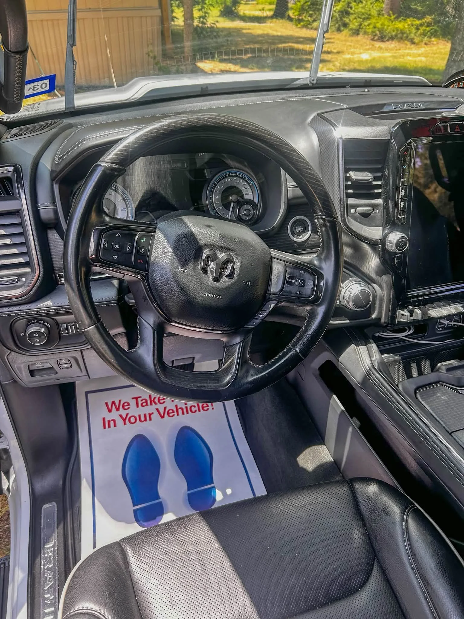 The interior of a vehicle shows the steering wheel, dashboard, air vents, and a large touchscreen display. A paper mat on the passenger side floor reads 'We Take... In Your Vehicle!' and features blue footprints.
