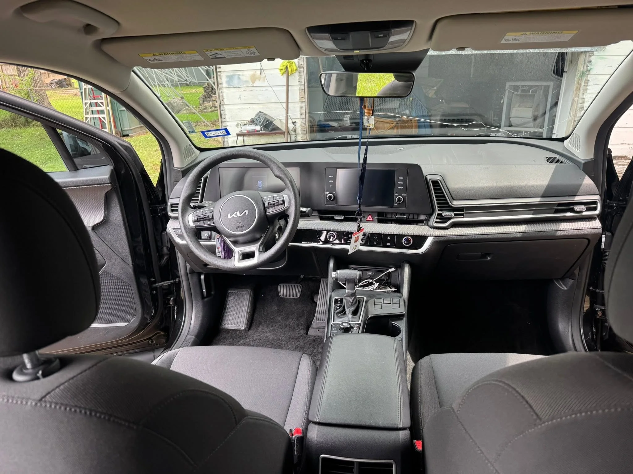 View from inside a Kia vehicle showing the dashboard, steering wheel, and front seats. The windshield reveals a house exterior with some equipment and outdoor items. The rearview mirror hangs from the center of the roof.
