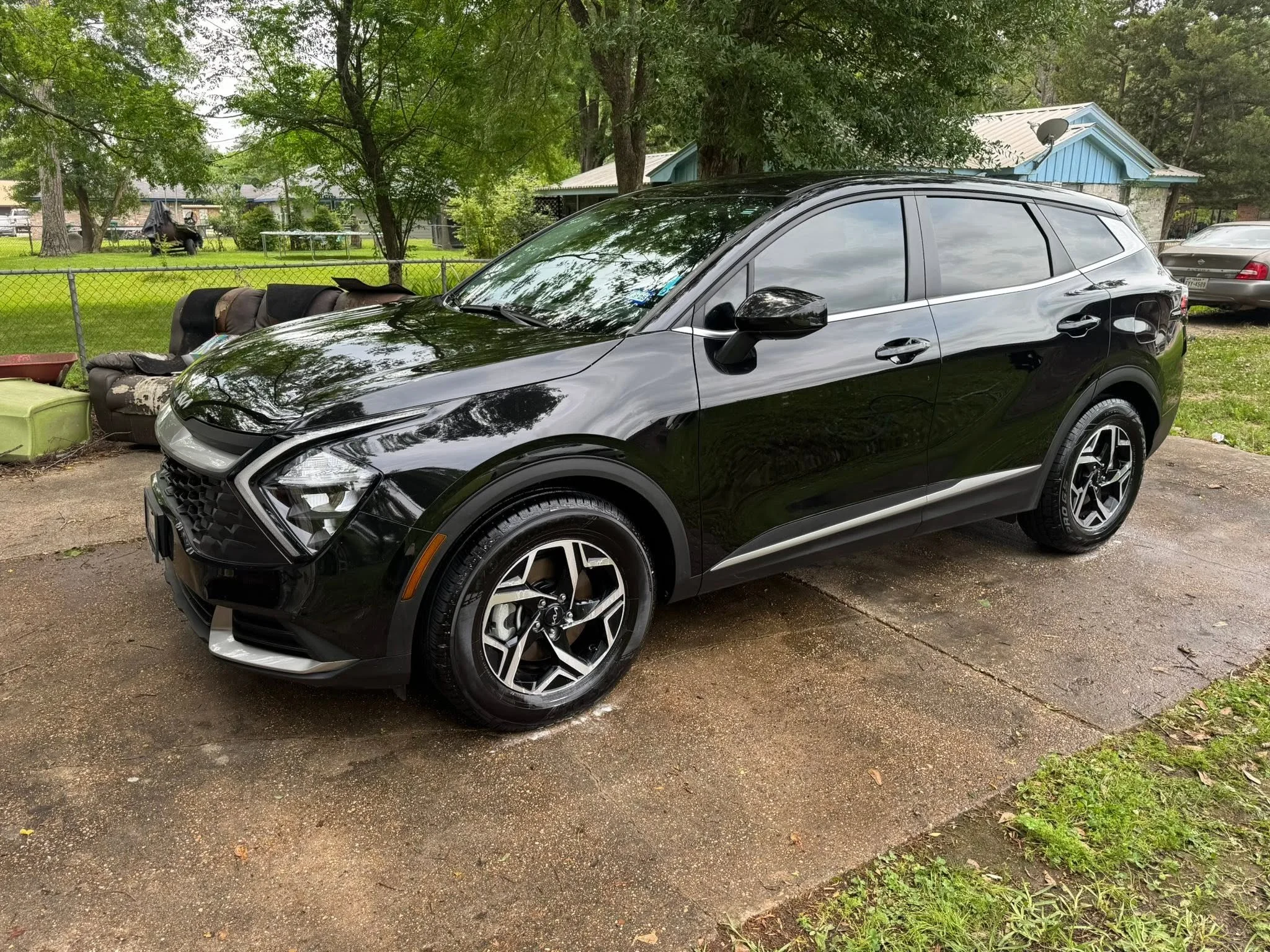 A black modern compact SUV parked in a backyard with a chain-link fence, trees, and a small house in the background.