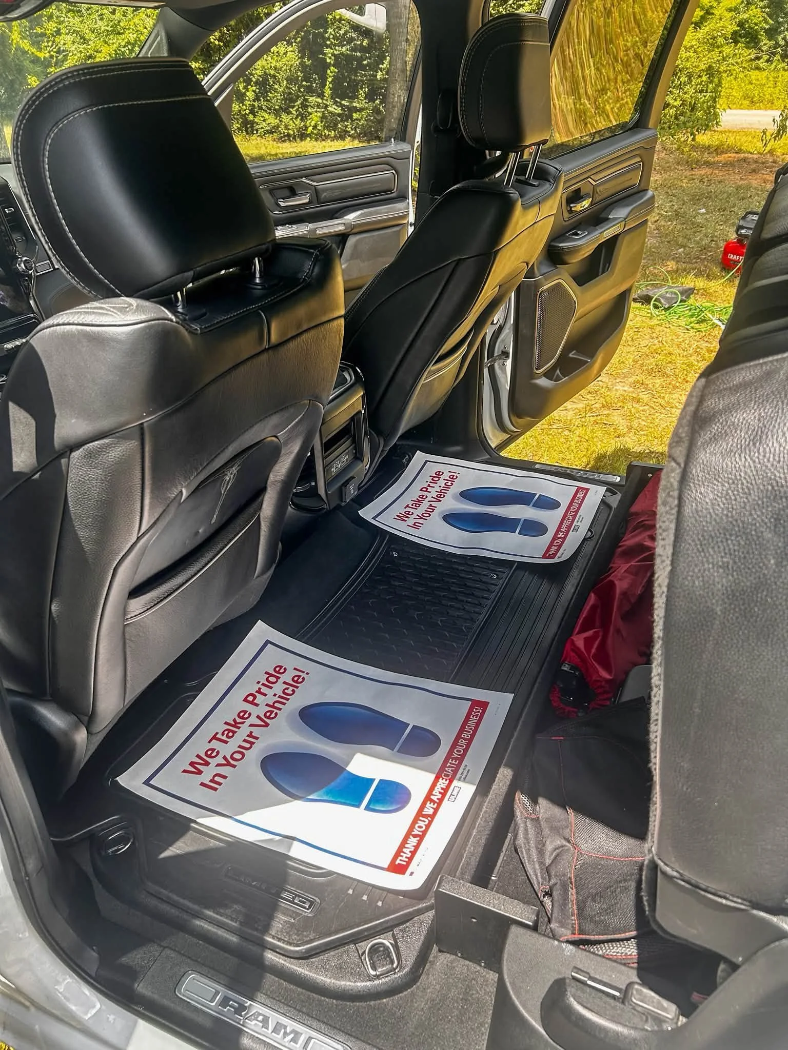 The interior of a vehicle showing black leather seats, some with headrests, and a floor mat with a sign that says "We Take Pride in Your Vehicle!" The door is open, and outside greenery is visible.