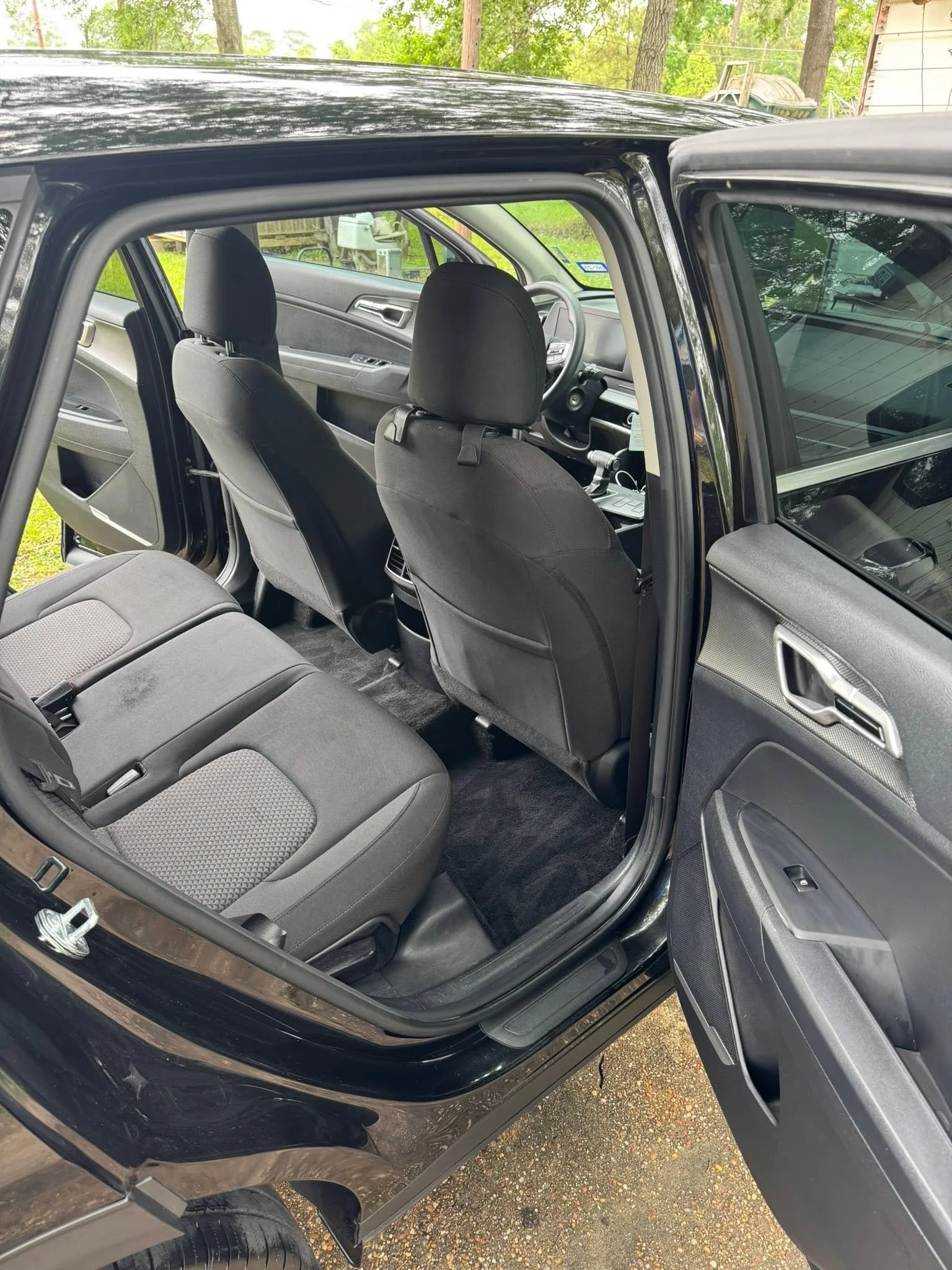 The interior of a black SUV showing the front and rear seats, dashboard, steering wheel, and open driver-side door with a view of a wooded yard outside.