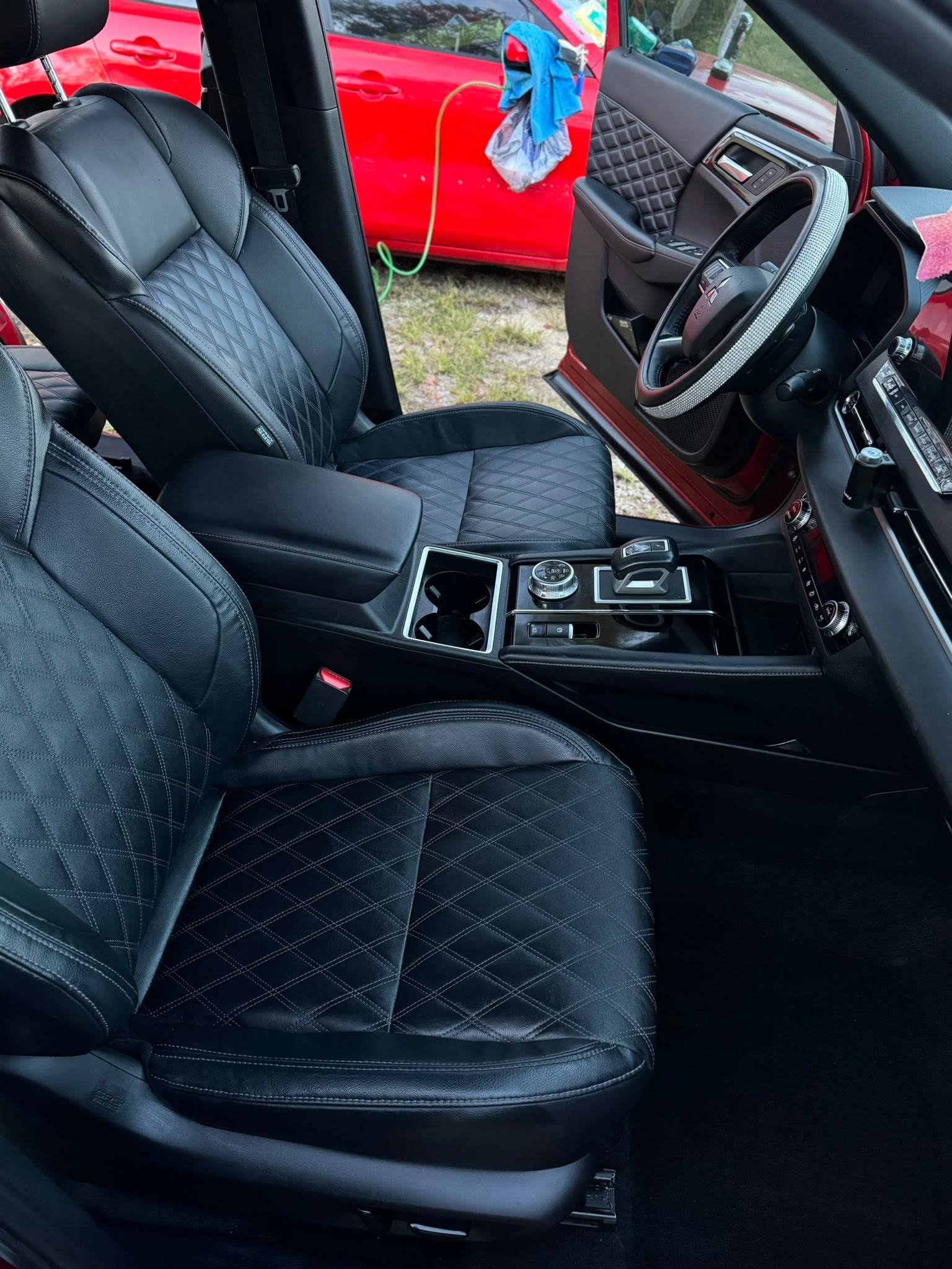 Inside view of a car showing black leather front seats with diamond quilted pattern, a black dashboard with silver accents, and a steering wheel with a silver and black cover. The passenger side door panel is also quilted. Outside the car, there is a