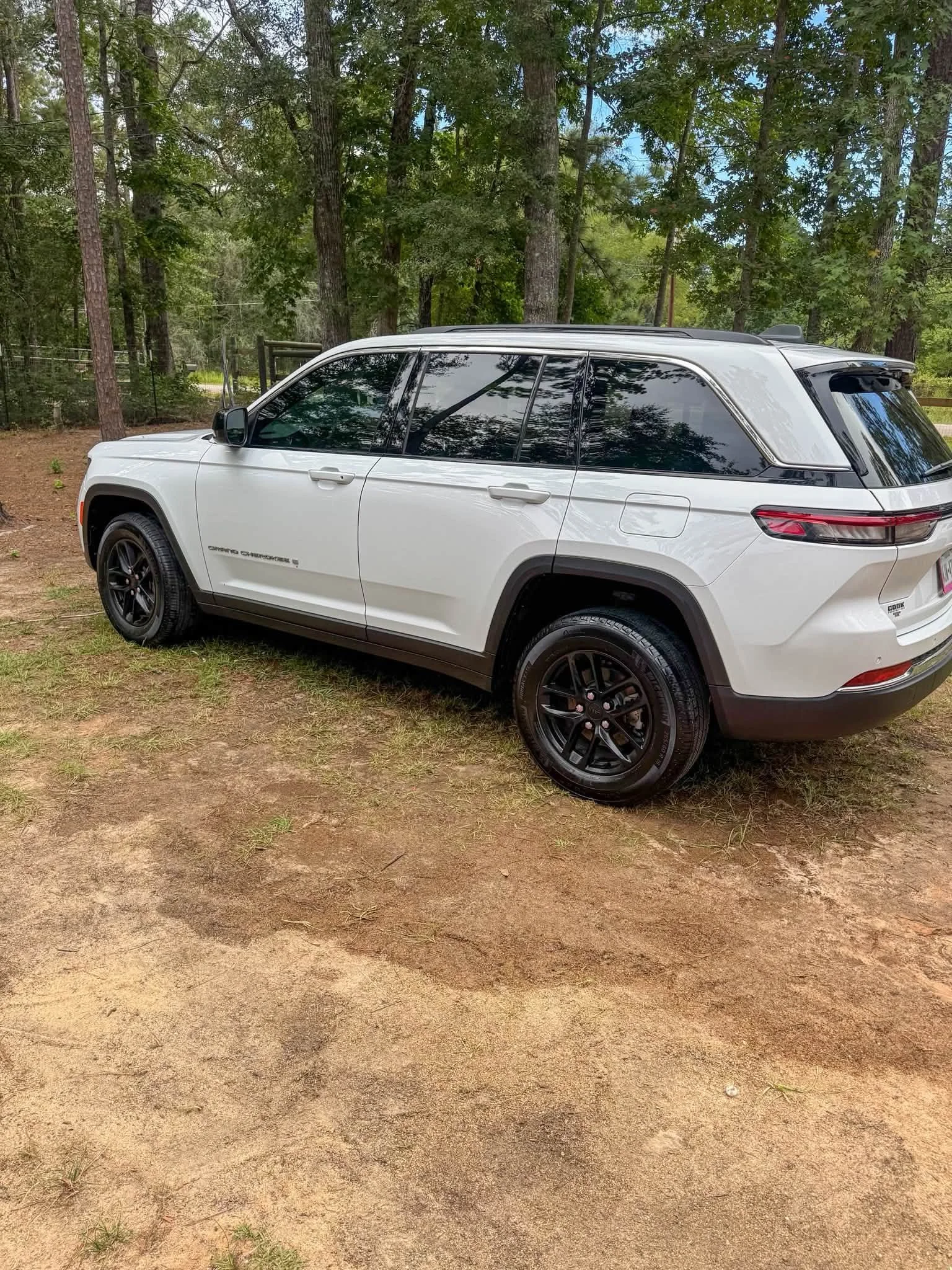 A white Jeep Grand Cherokee parked on a dirt surface with trees and a fence in the background.
