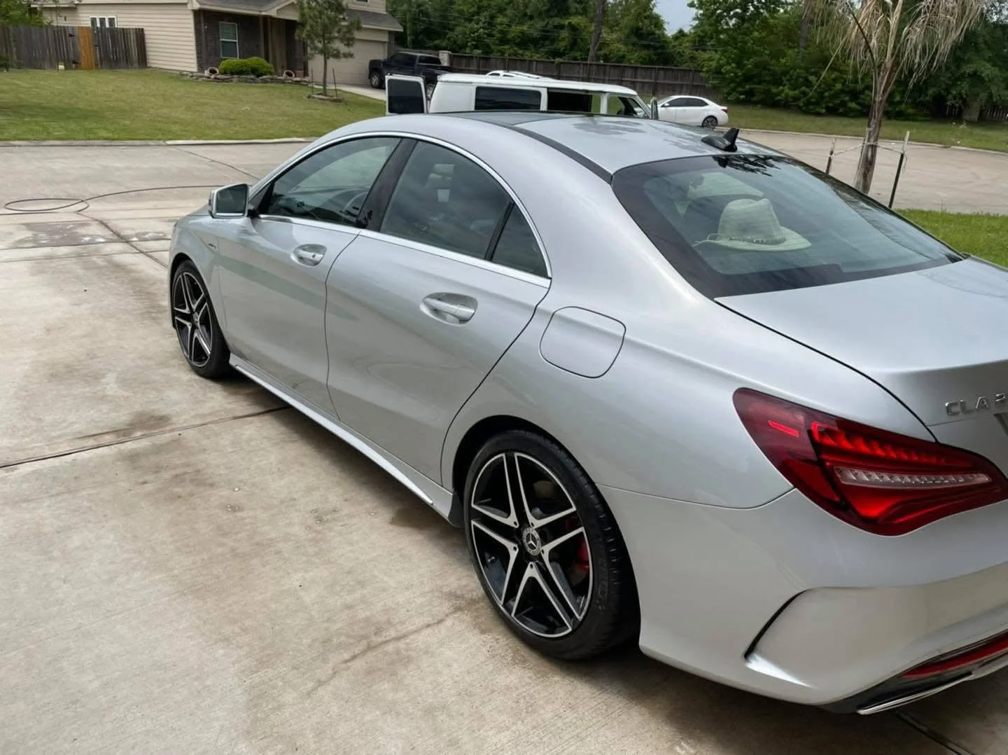 Silver Mercedes-Benz CLA parked on a concrete driveway in a residential area, with a grassy lawn, trees, houses, and other vehicles in the background.