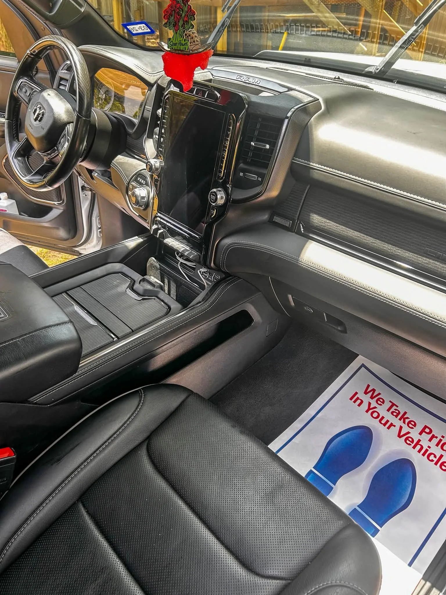 Interior of a vehicle showing the dashboard, steering wheel, and front seats. There is a red and green decorative item hanging from the rearview mirror, and a sign on the floor that reads, 'We Take Pride in Your Vehicle.'