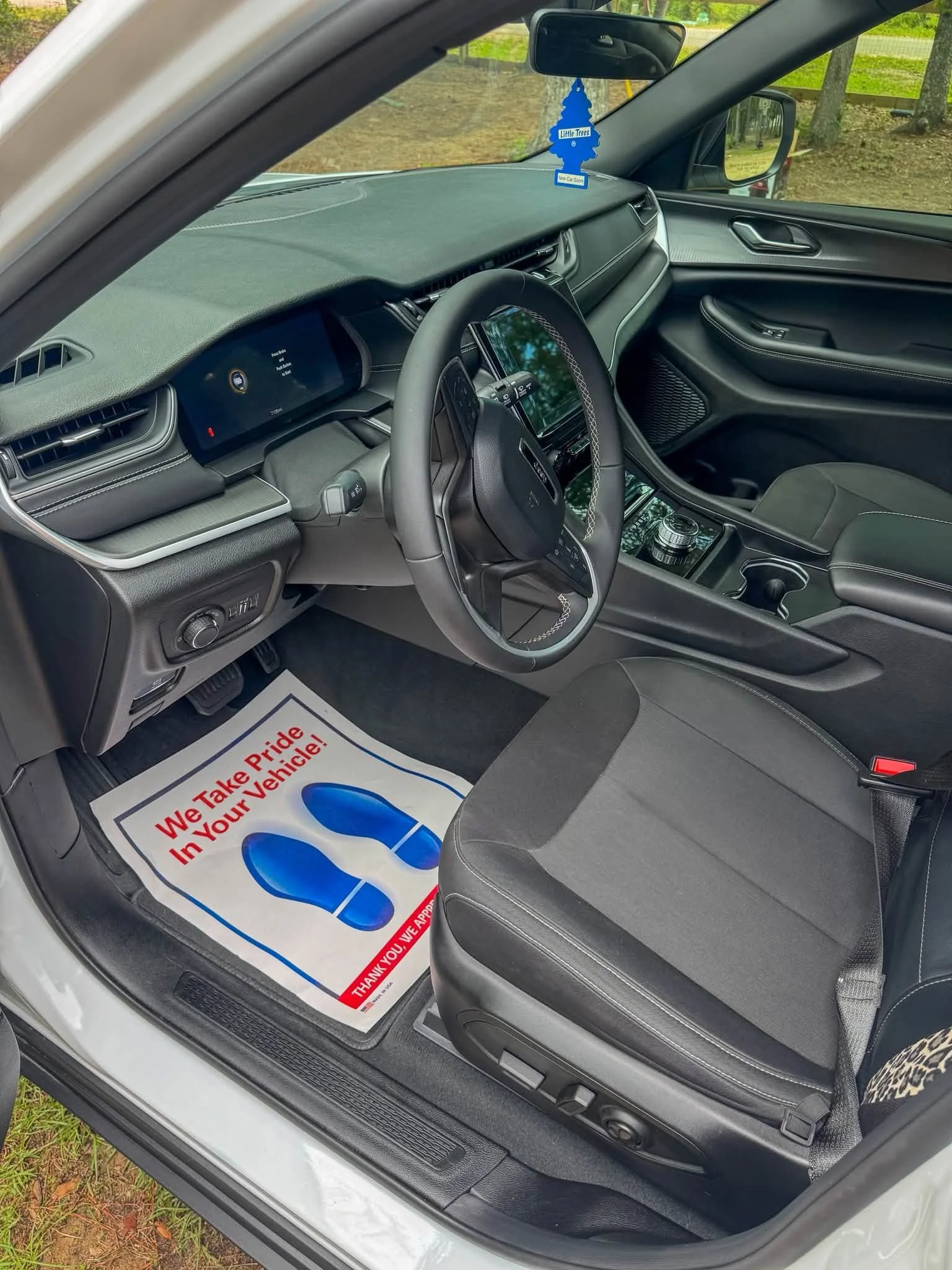 Interior of a car showing the driver's side, steering wheel, dashboard, and a floor mat with a message that says "We Take Pride in Your Vehicle!" in red and blue lettering.