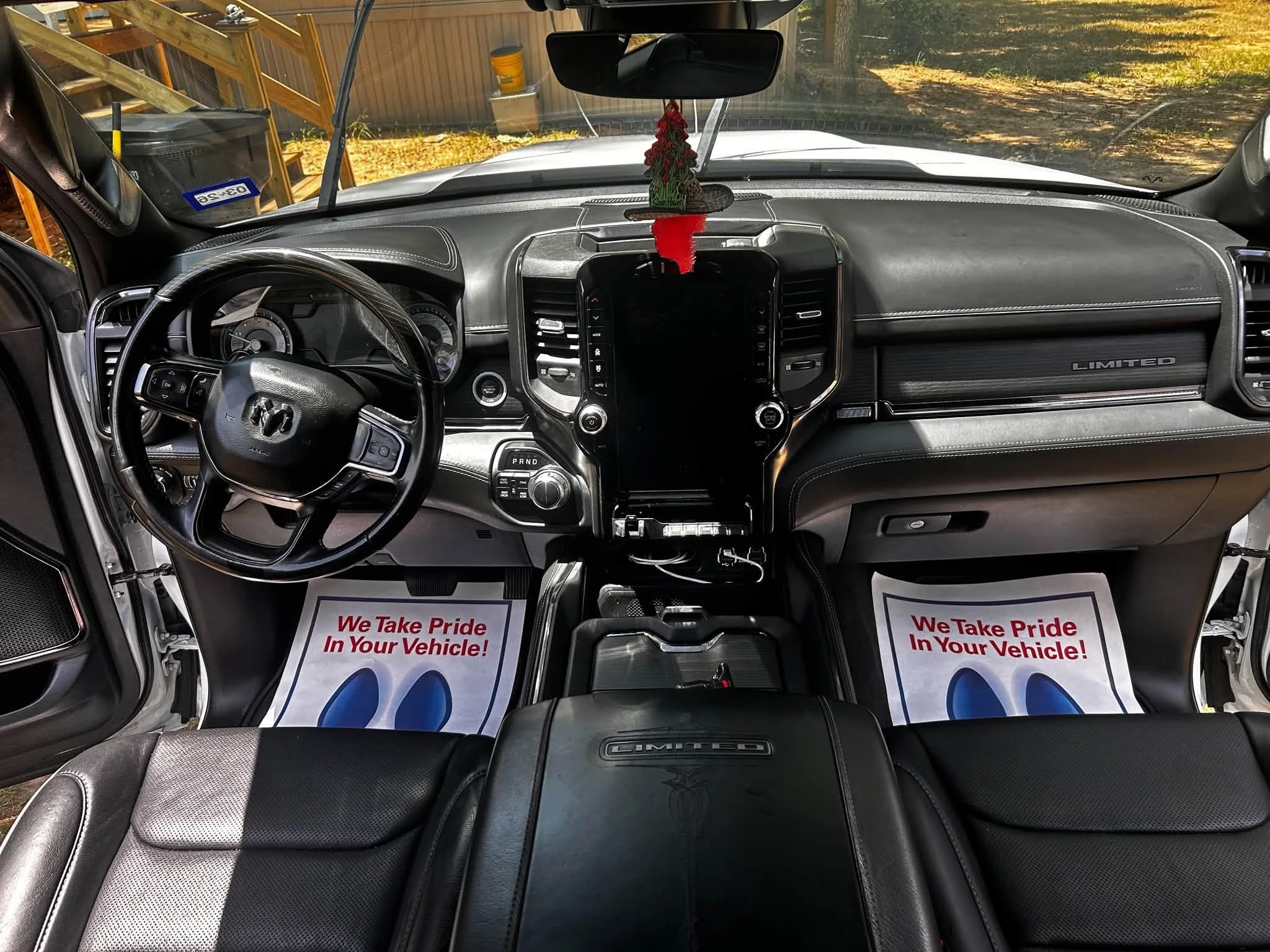Interior view of a vehicle showing the dashboard, steering wheel, and front seats. There are two paper mats on the floor with the text "We Take Pride In Your Vehicle!". In the background, the weather is sunny outside.