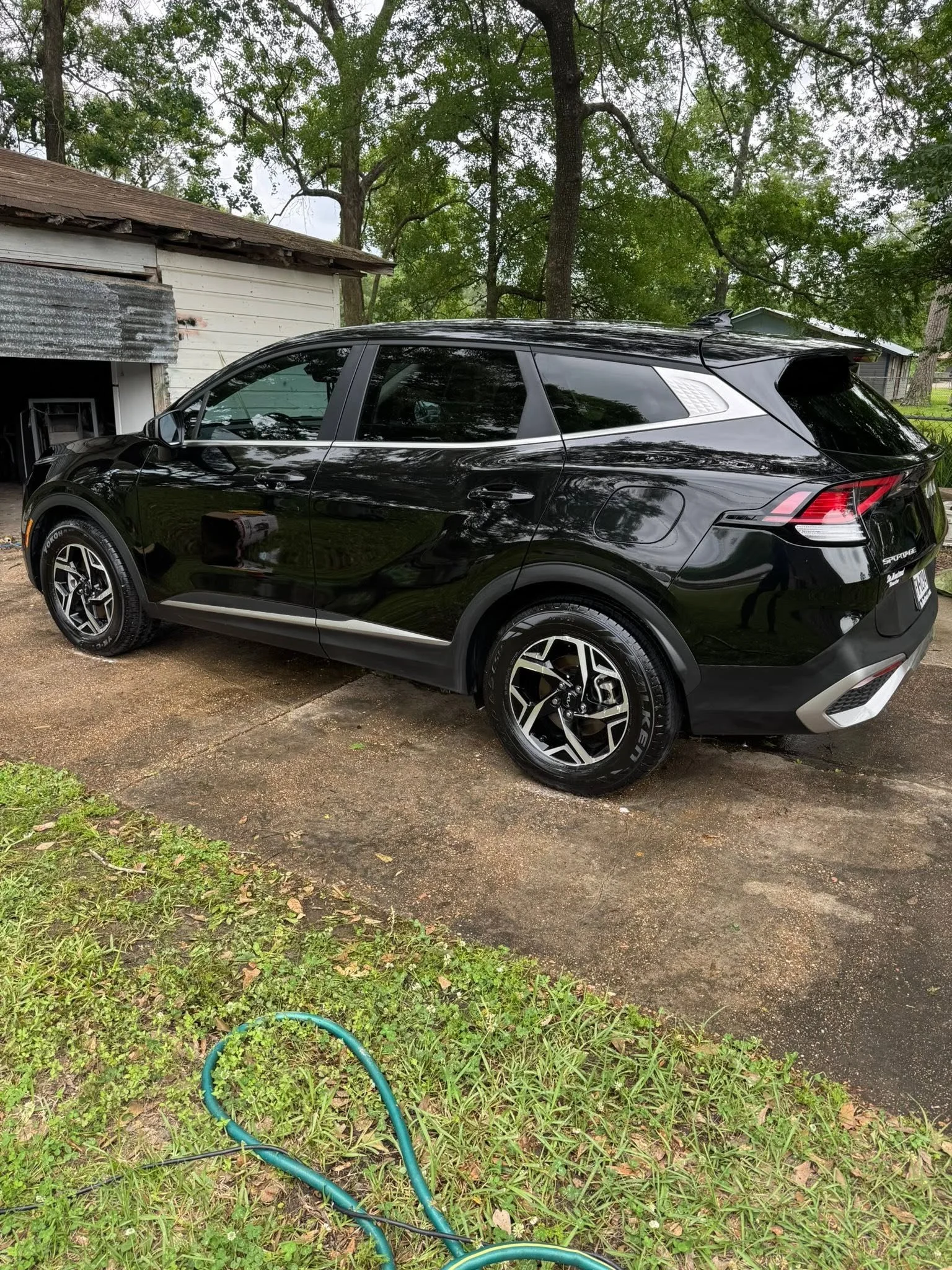 A black SUV car parked on a driveway next to a white and weathered shed, with trees and a house in the background, and a green garden hose on the grass in the foreground.
