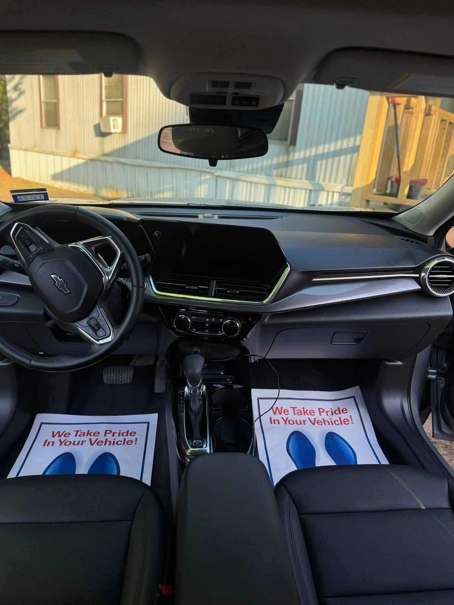 Inside view of a Chevrolet vehicle showing the dashboard, steering wheel, rearview mirror, and two paper floor mats with the message "We Take Pride In Your Vehicle!" on the front seats.