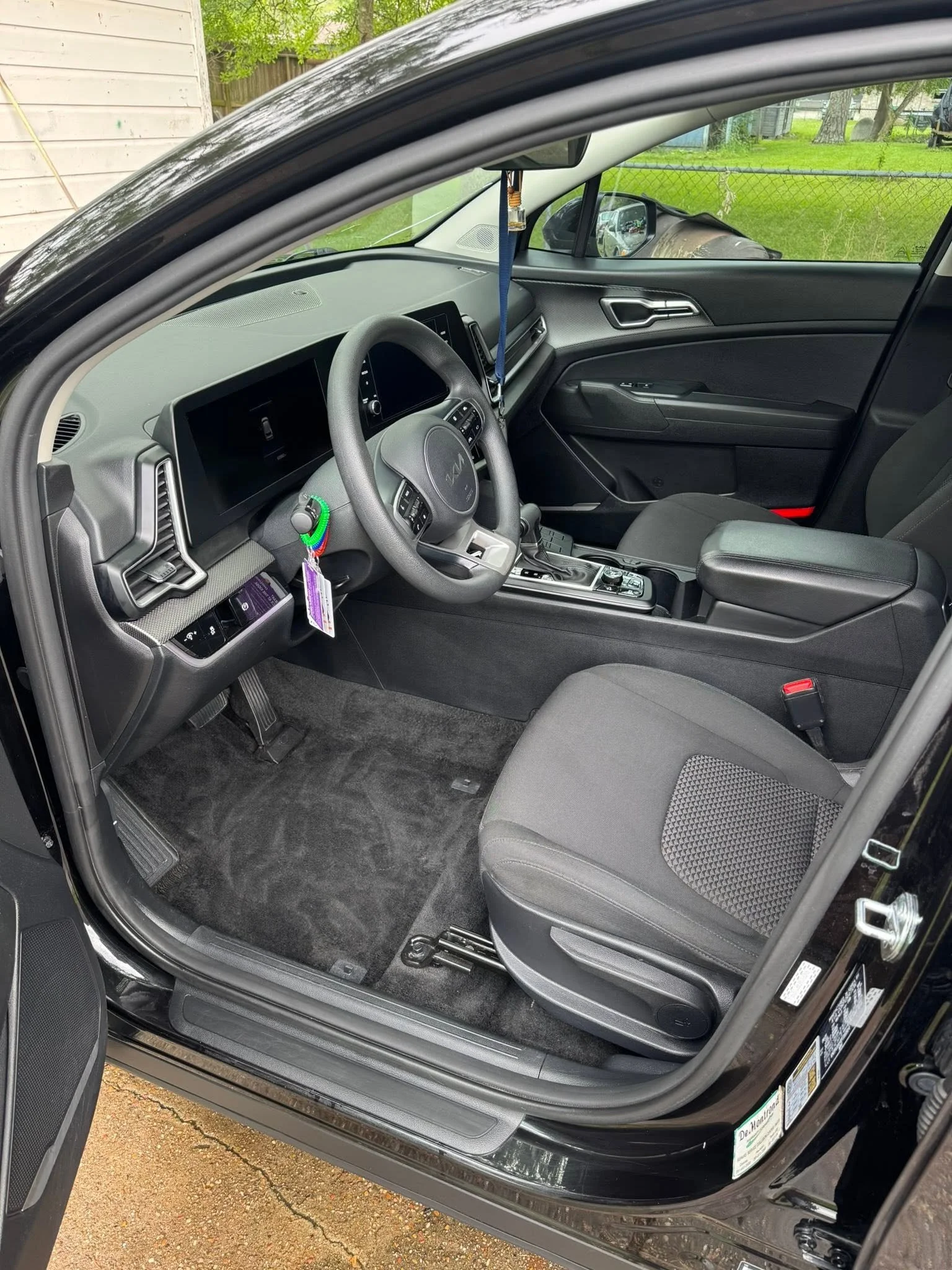 The interior of a black modern car, showing the driver's seat, steering wheel, dashboard, and part of the front passenger seat, with greenery and a fence visible outside through the windows.