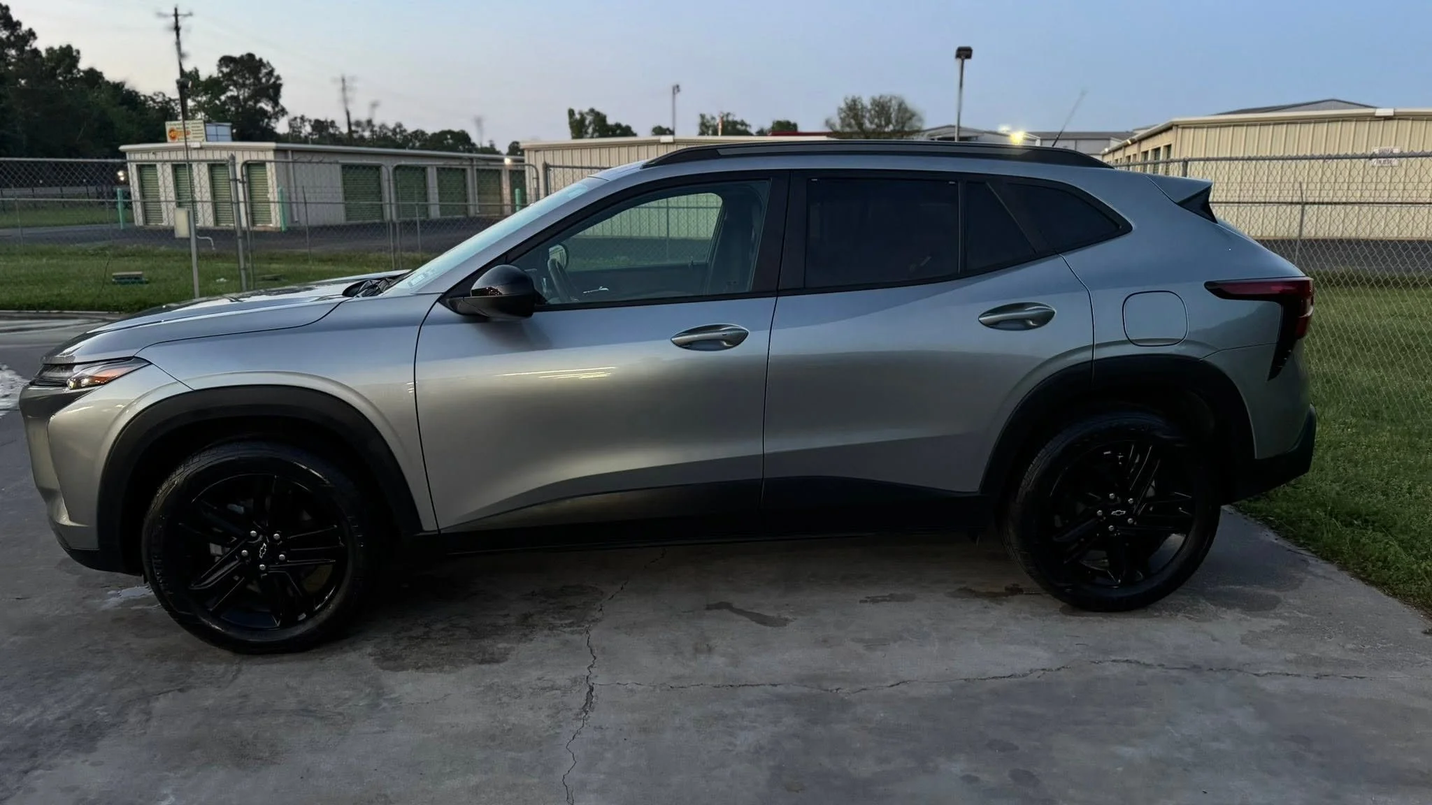 Silver compact SUV parked on concrete lot near a chain-link fence with trees and buildings in the background.
