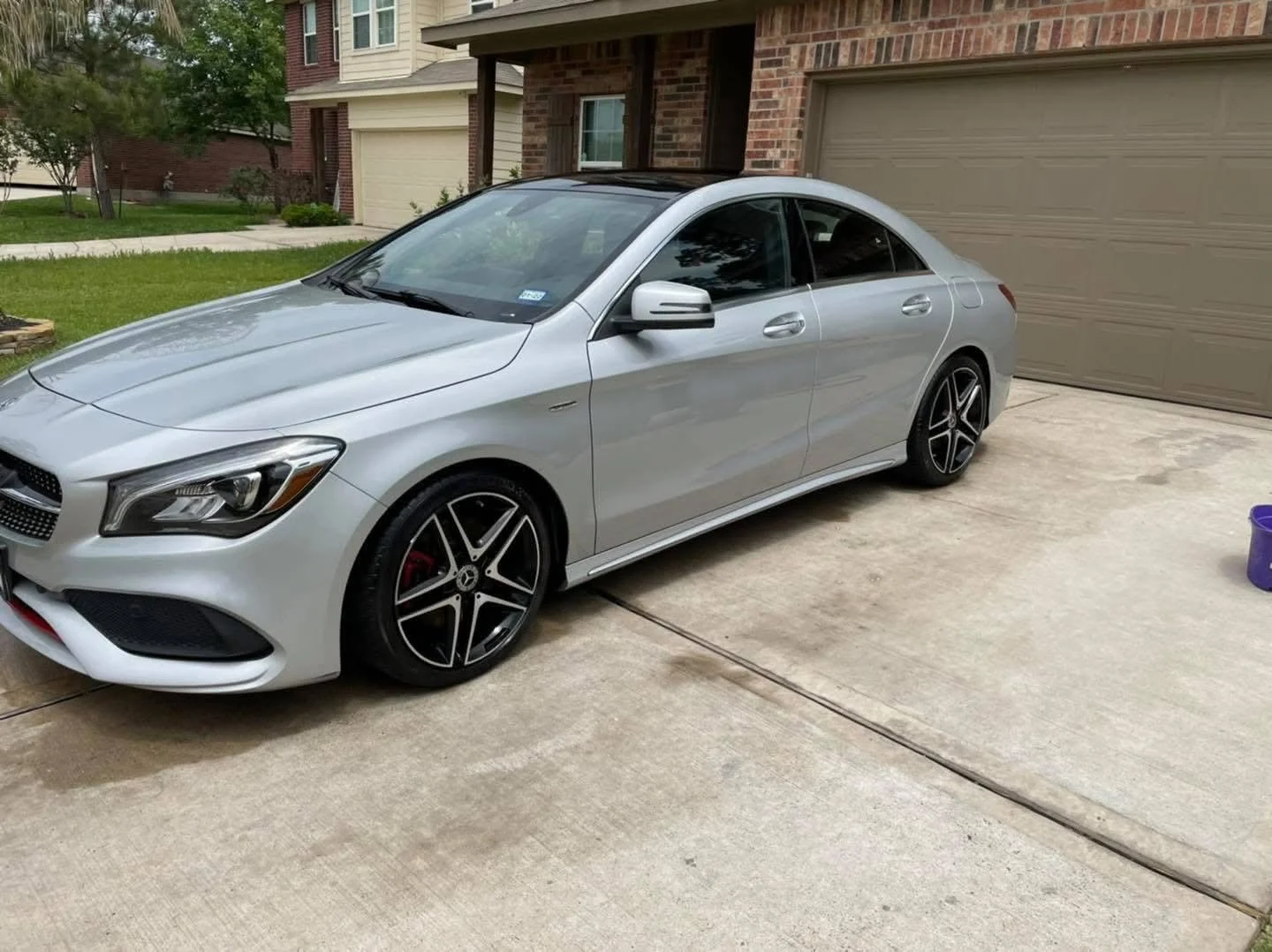 Silver luxury sedan parked in driveway next to a closed garage door, with a brick house and front lawn in the background.