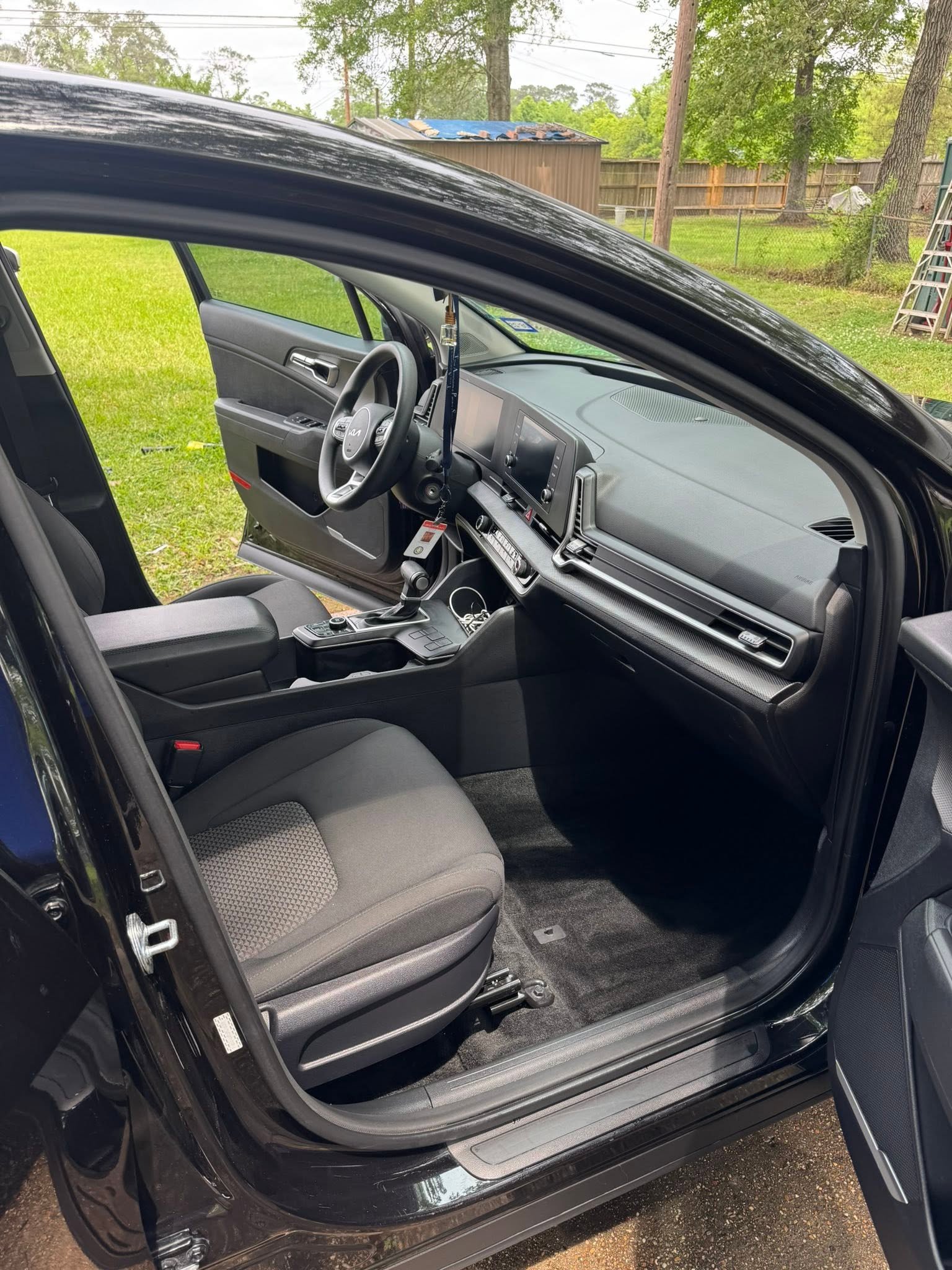The interior of a black car showing the front passenger seat, dashboard, steering wheel, and center console with a background of a backyard with trees, a shed, and a wooden fence.