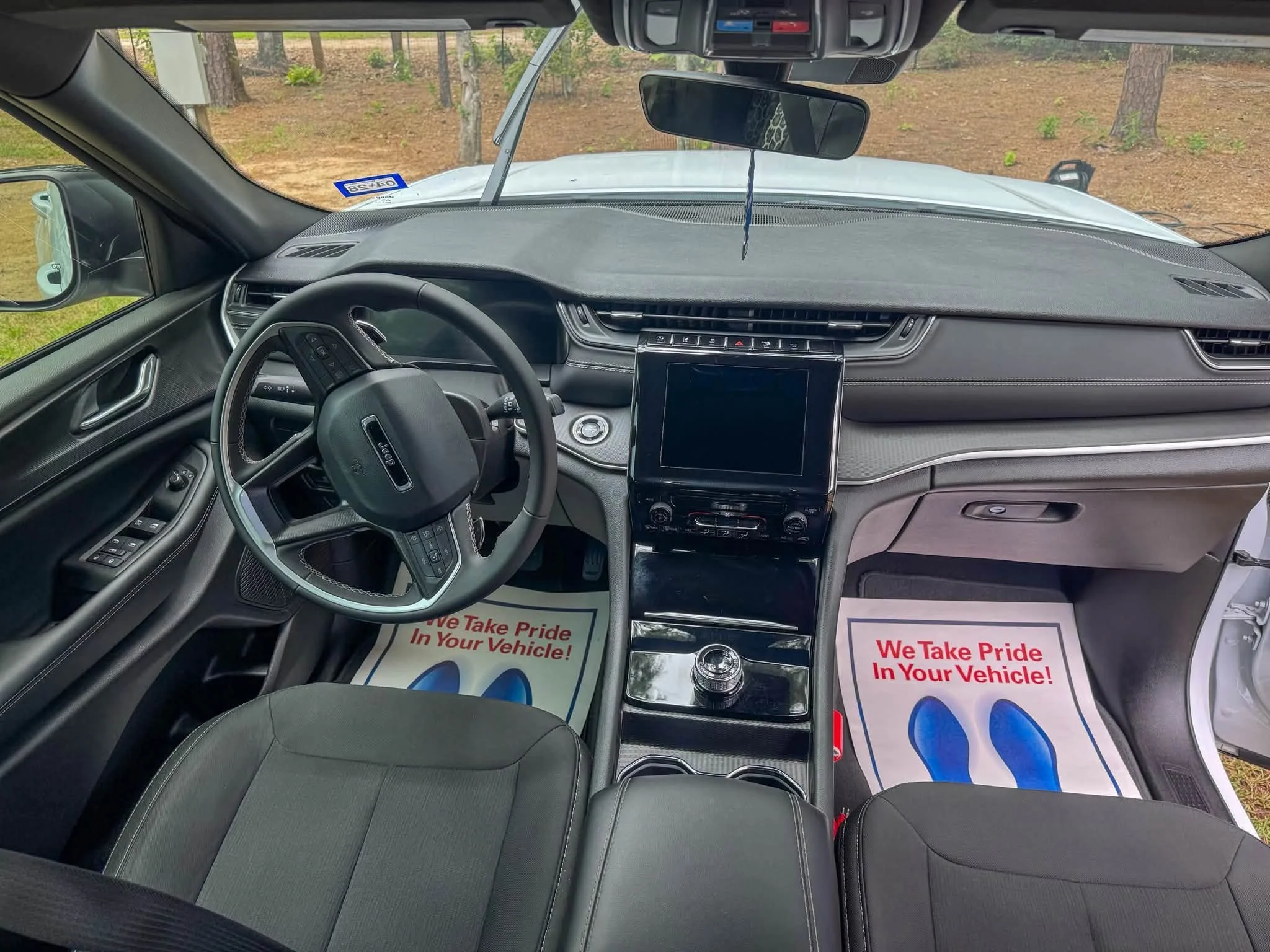 View from inside a vehicle showing the dashboard, steering wheel, and front seats. Outside, there are trees visible through the windshield, and a sign is attached to the windshield. There are also two paper floor mats with red text that says, 'We Tak