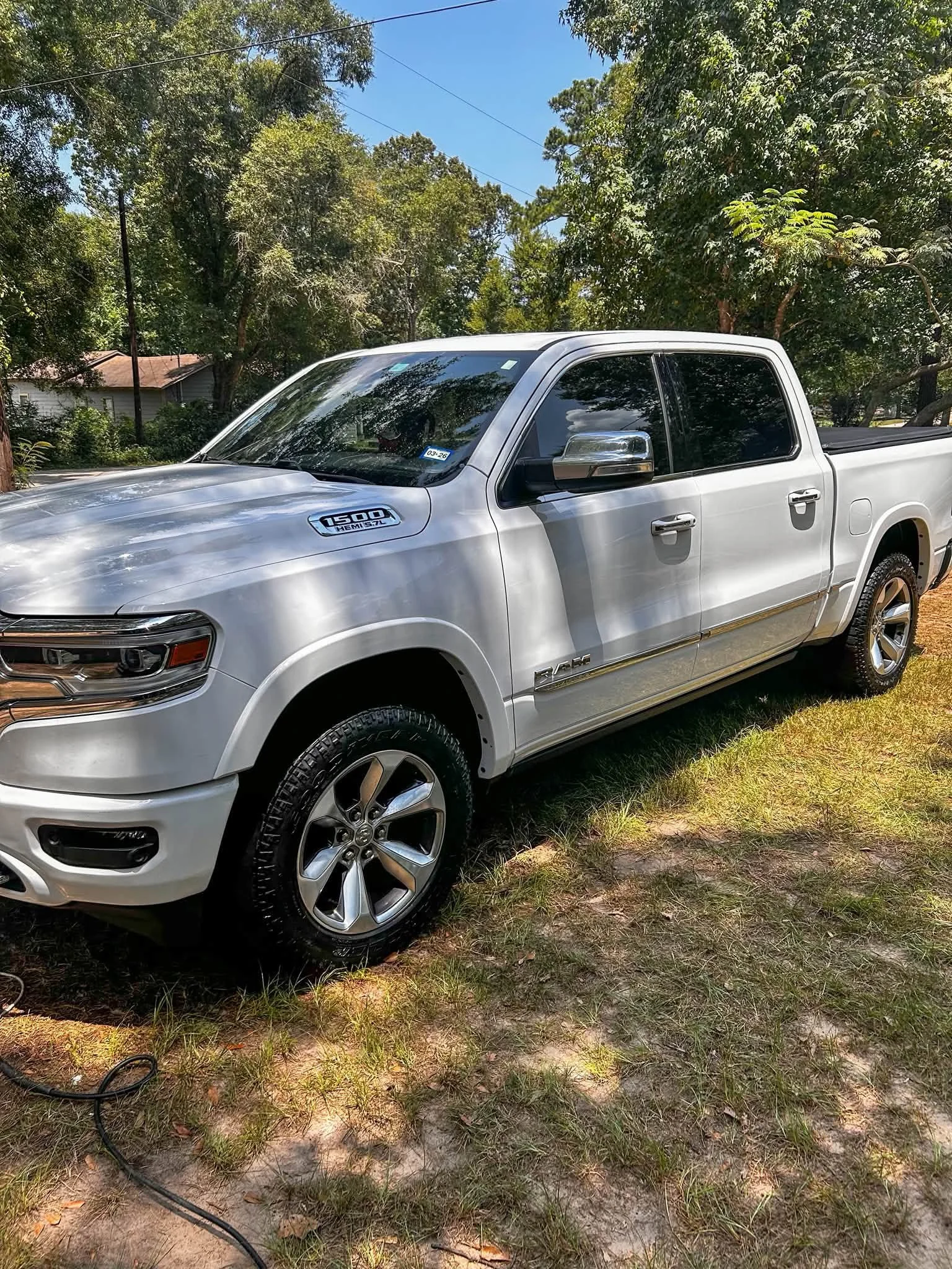 A silver pickup truck parked on grass and dirt under trees with a house and power lines in the background.