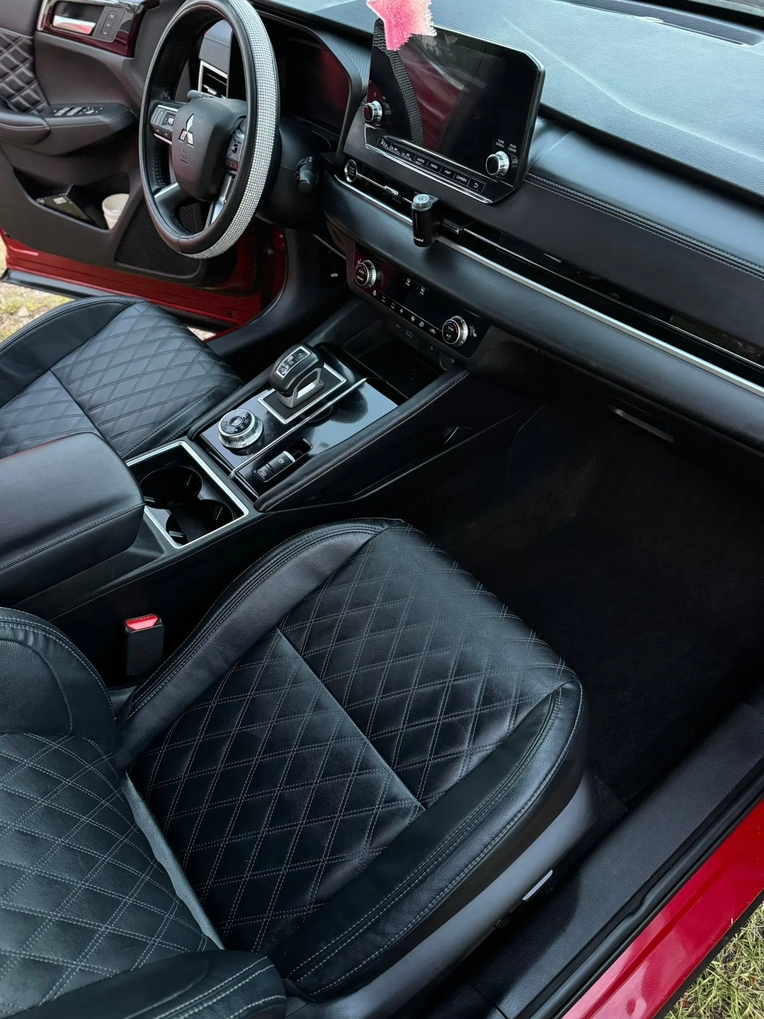 The interior of a car showing black quilted leather seats, a gear shift, a steering wheel with a diamond-patterned cover, a dashboard with a large central display, and various controls and buttons.