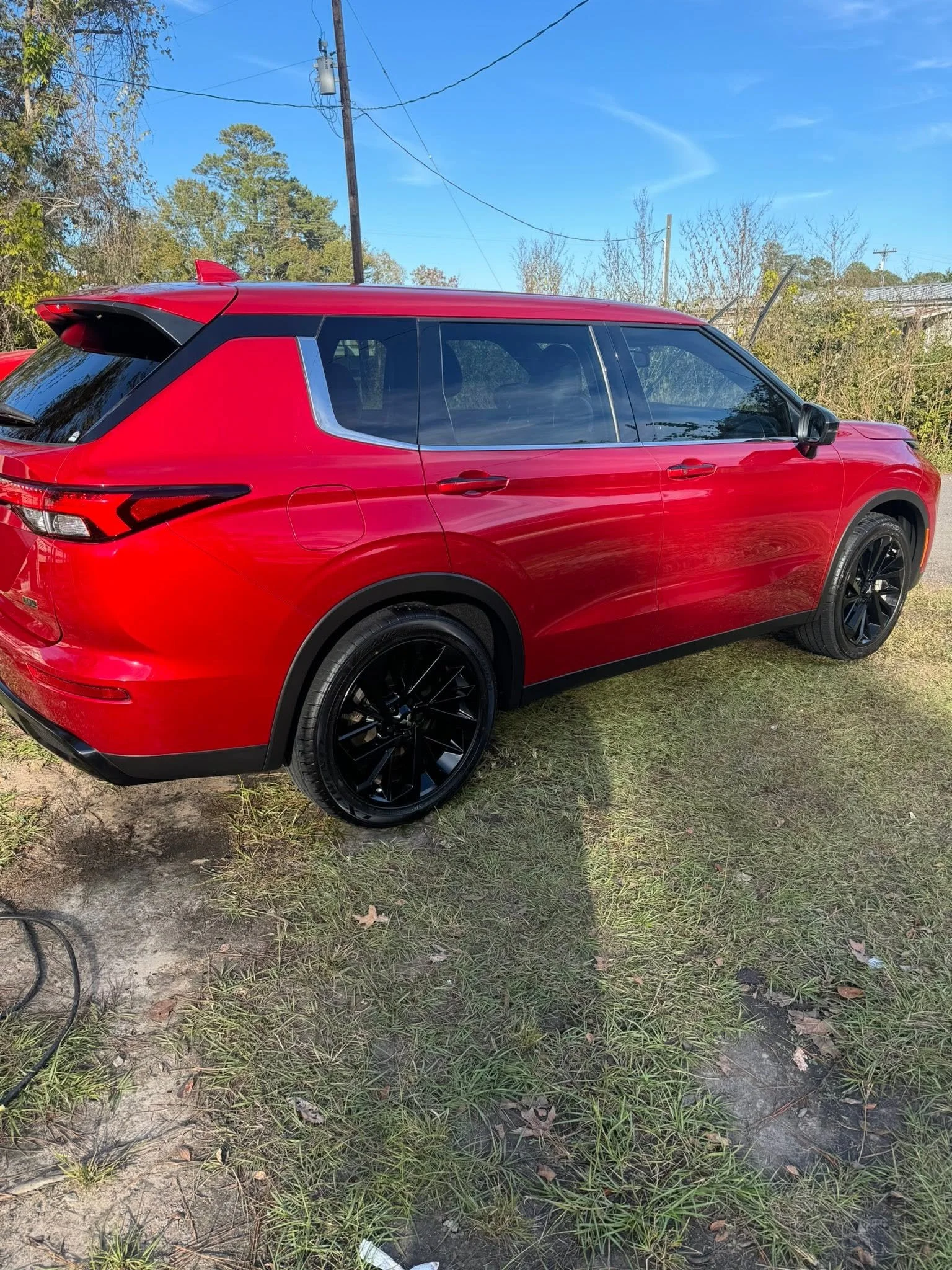 Red SUV parked on grass and dirt ground, background includes trees, utility poles, and a clear blue sky.