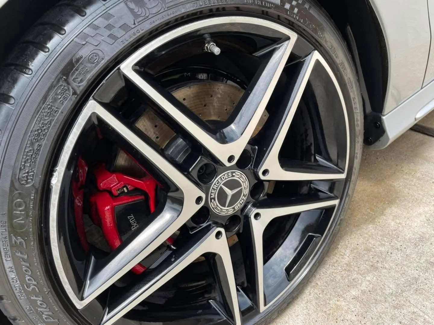 Close-up of a Mercedes-Benz wheel with a black and silver alloy rim, featuring a red brake caliper and a visible brake disc, on a concrete surface.
