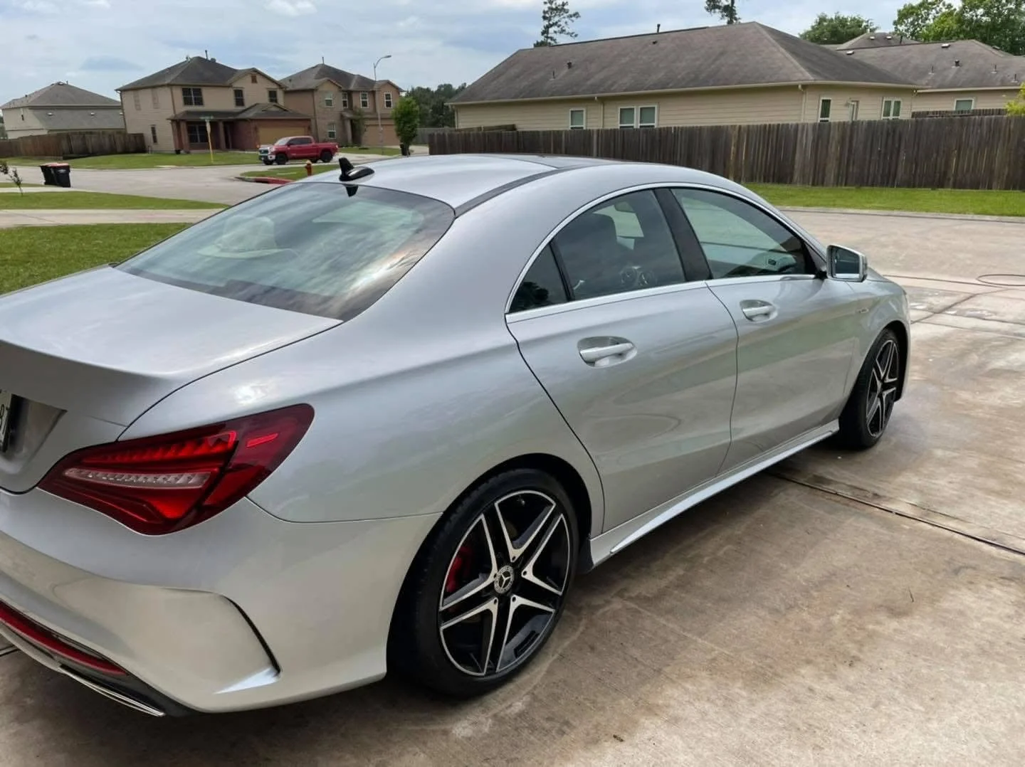 A silver luxury sedan parked on a concrete driveway in a suburban neighborhood surrounded by houses, fences, and a grassy yard.