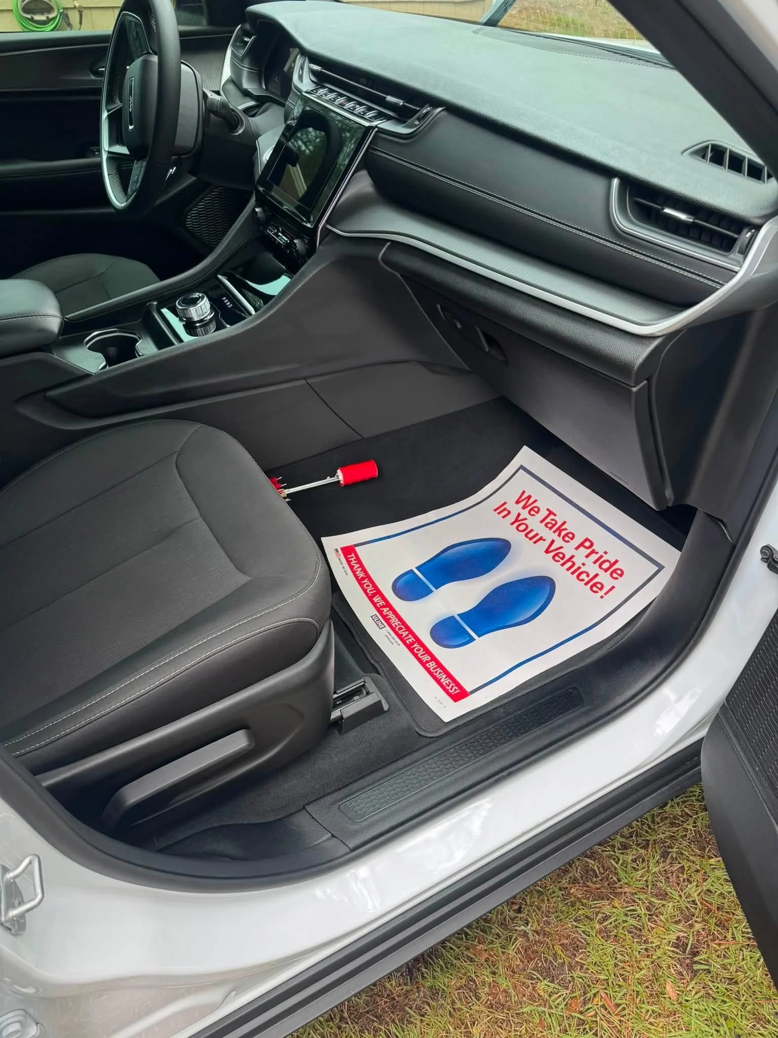 Front interior of a modern vehicle, showing the dashboard, steering wheel, center console with controls, and driver's seat. A paper floor mat with a blue footprint and red text reading "We Take Pride in Your Vehicle!" is on the floor.