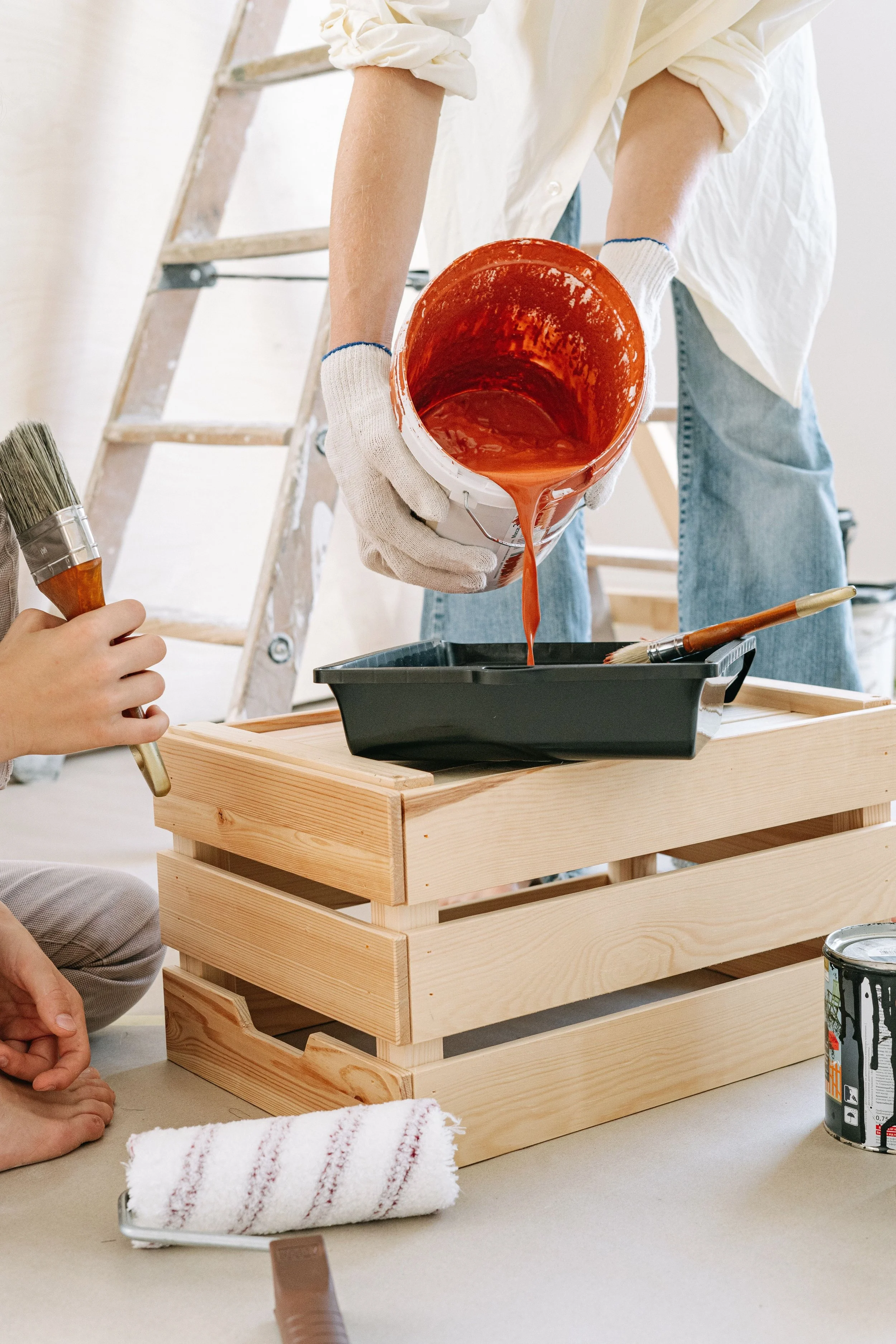 Person pouring orange paint from a bucket onto a black tray, with a wooden step ladder in the background, a paintbrush and a can of black spray paint visible on a table, and someone sitting nearby holding a paintbrush.