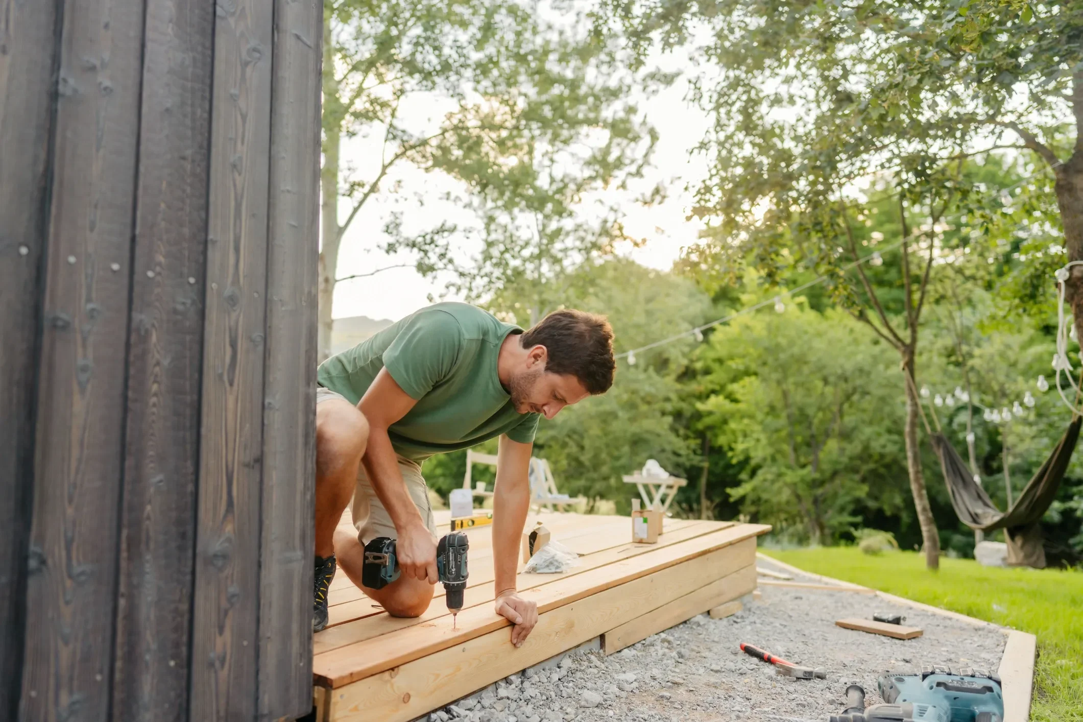A young man wearing a green t-shirt working outdoors on a woodworking project, using a cordless drill on a wooden deck with trees and string lights in the background.