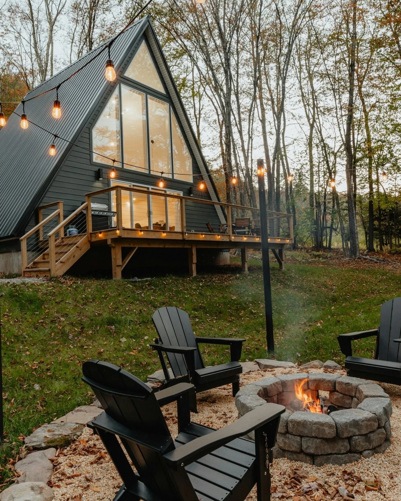 A modern A-frame house with large windows and a wooden deck, decorated with string lights, surrounded by trees in autumn. In the foreground, there are black Adirondack chairs arranged around a small fire pit with glowing fire. The scene is set in the evening or early night.