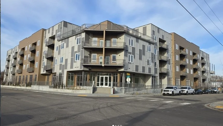 Modern multi-story apartment building with balconies, large windows, and a combination of gray, brown, and beige exterior siding, situated on a city street with parked cars.