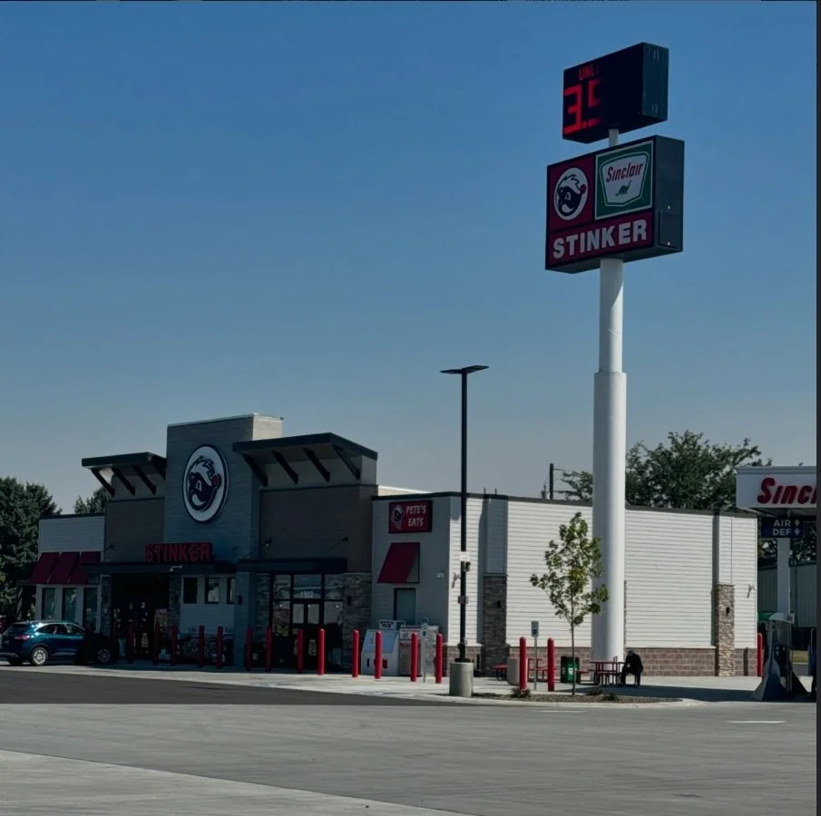 A fast-food restaurant called Stinker with a large sign displaying the number 36. The building has a white exterior with a logo featuring a stylized face. There are red bollards in front of the entrance, and a parking lot with a few cars. The scene i