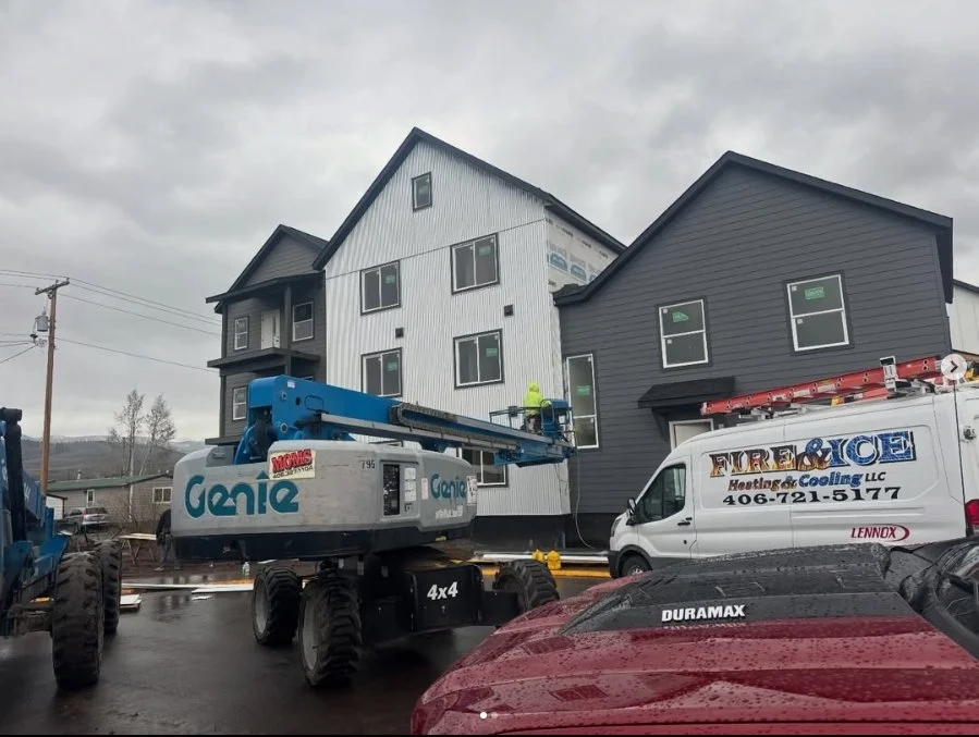 Construction workers installing exterior siding on a multi-story residential building using a blue boom lift. A white van labeled 'FIRE & ICE Heating & Cooling LLC' is parked nearby on a wet road, with several other vehicles around.