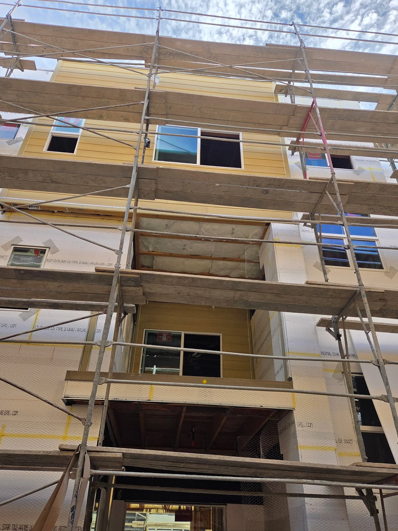 Construction scaffolding surrounding a building under construction with yellow exterior siding and some windows, set against a partly cloudy sky.