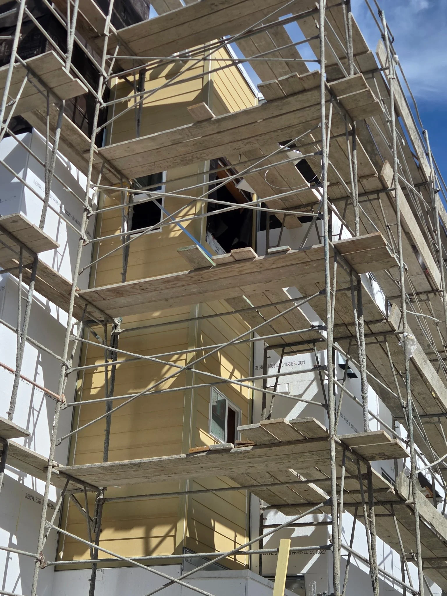 Construction scaffolding surrounds a yellow two-story house under construction. The scaffolding includes multiple wooden planks and metal poles. The house has large windows and some parts of the exterior walls are visible. Blue sky and sunlight are a