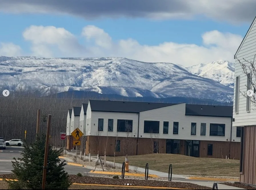 Residential buildings with white siding and black roofs, mountain range with snow in the background, a pedestrian crossing sign, and a stop sign in the foreground.