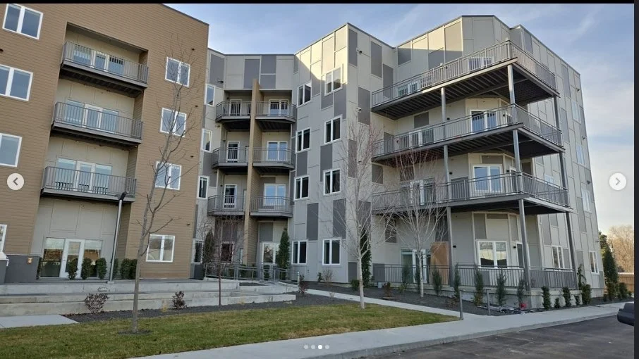 Modern multi-story residential apartment building with balconies, surrounded by trees and a parking lot.