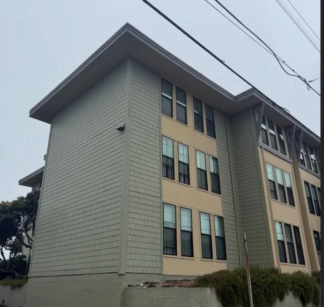 Photo of a multi-story building with beige walls and multiple windows, taken from below. Overhead power lines are visible at the top of the image.
