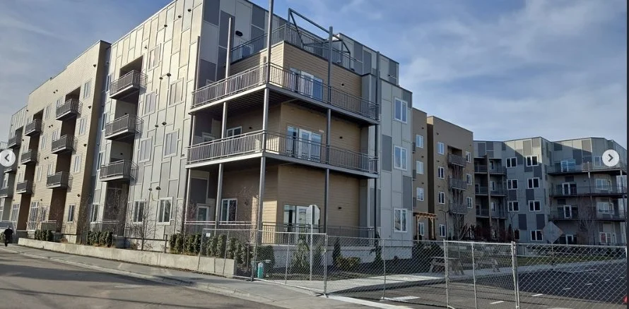 Modern multi-story apartment building with balconies, windows, and a fenced perimeter, under a partly cloudy sky.