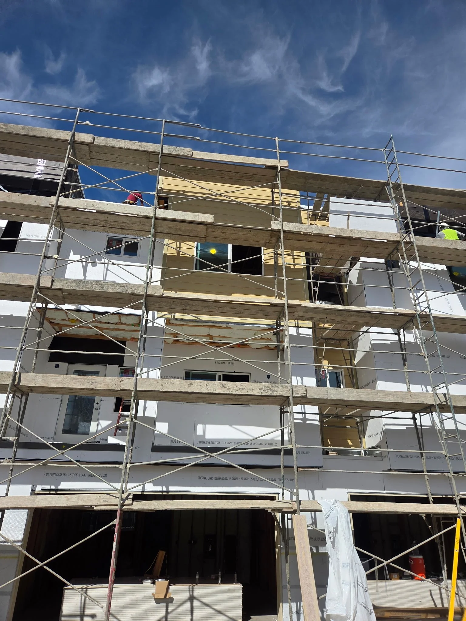 Construction site with scaffolding, workers, and a building under construction against a blue sky with clouds.