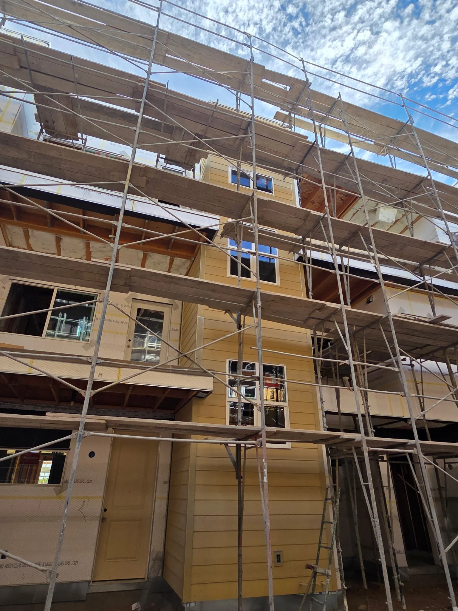 View of a multi-story house under construction with yellow siding, surrounded by scaffolding, against a blue sky with clouds.