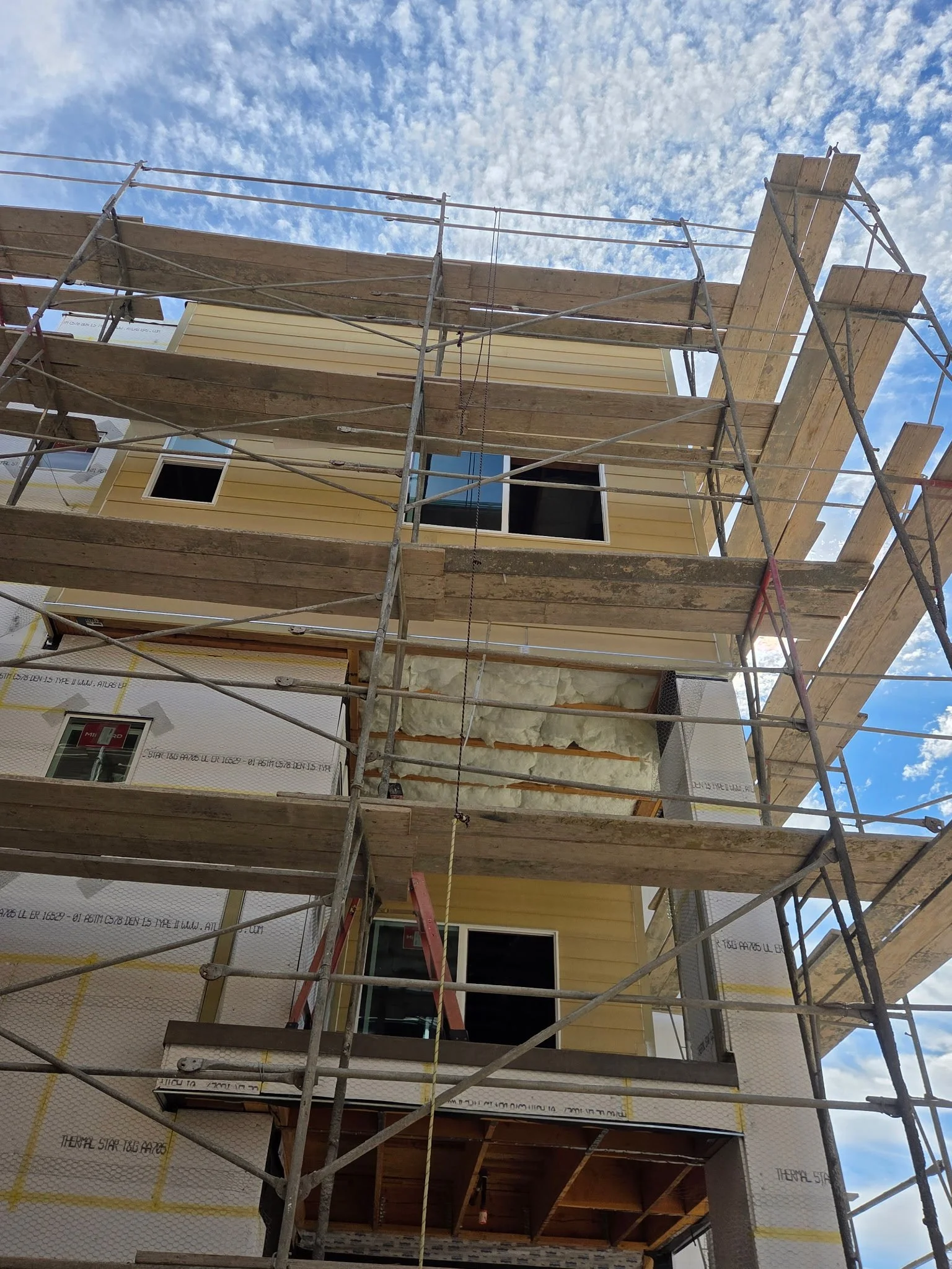 A multi-story building under construction with wooden framework and insulation, surrounded by metal scaffolding, against a background of a partly cloudy sky.