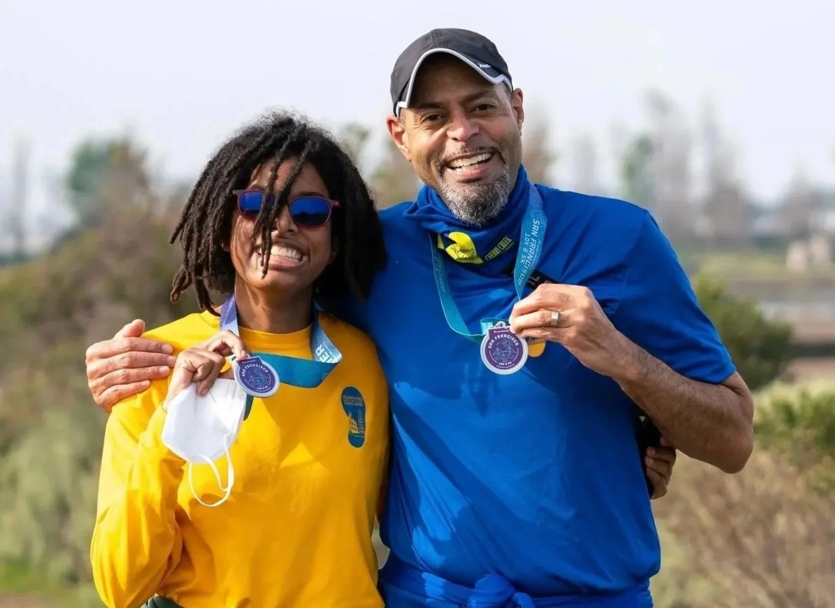 Two runners smiling with medals outdoors, one woman in a yellow shirt and sunglasses, and one man in a blue shirt with a cap, standing close together.
